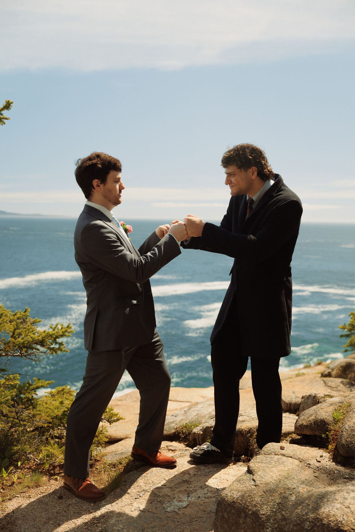 Two grooms exchange rings on rocky coastal cliffs during an outdoor wedding ceremony.