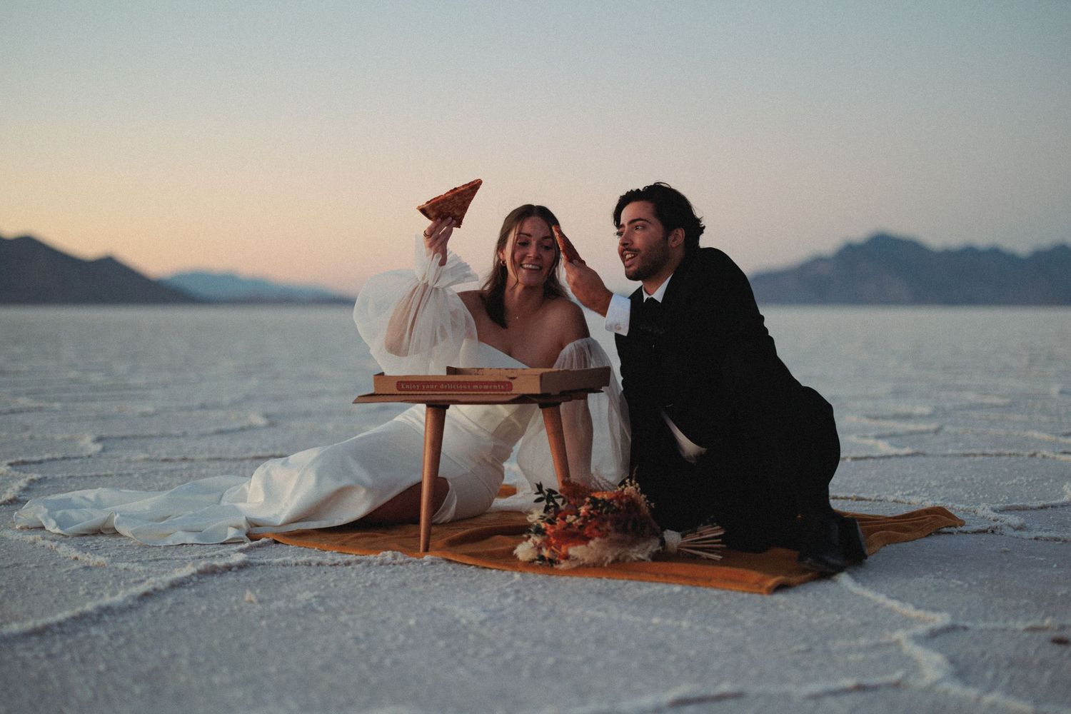 Couple relaxing on salt flats during sunset with mountains in the background.