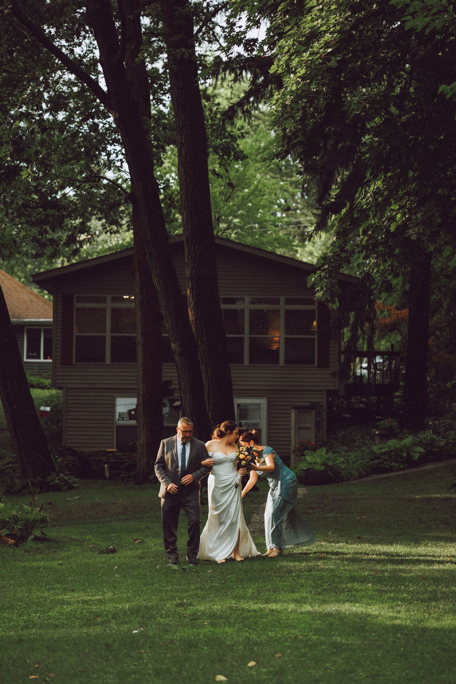 Wedding party walking across a green lawn with wooden house in background.
