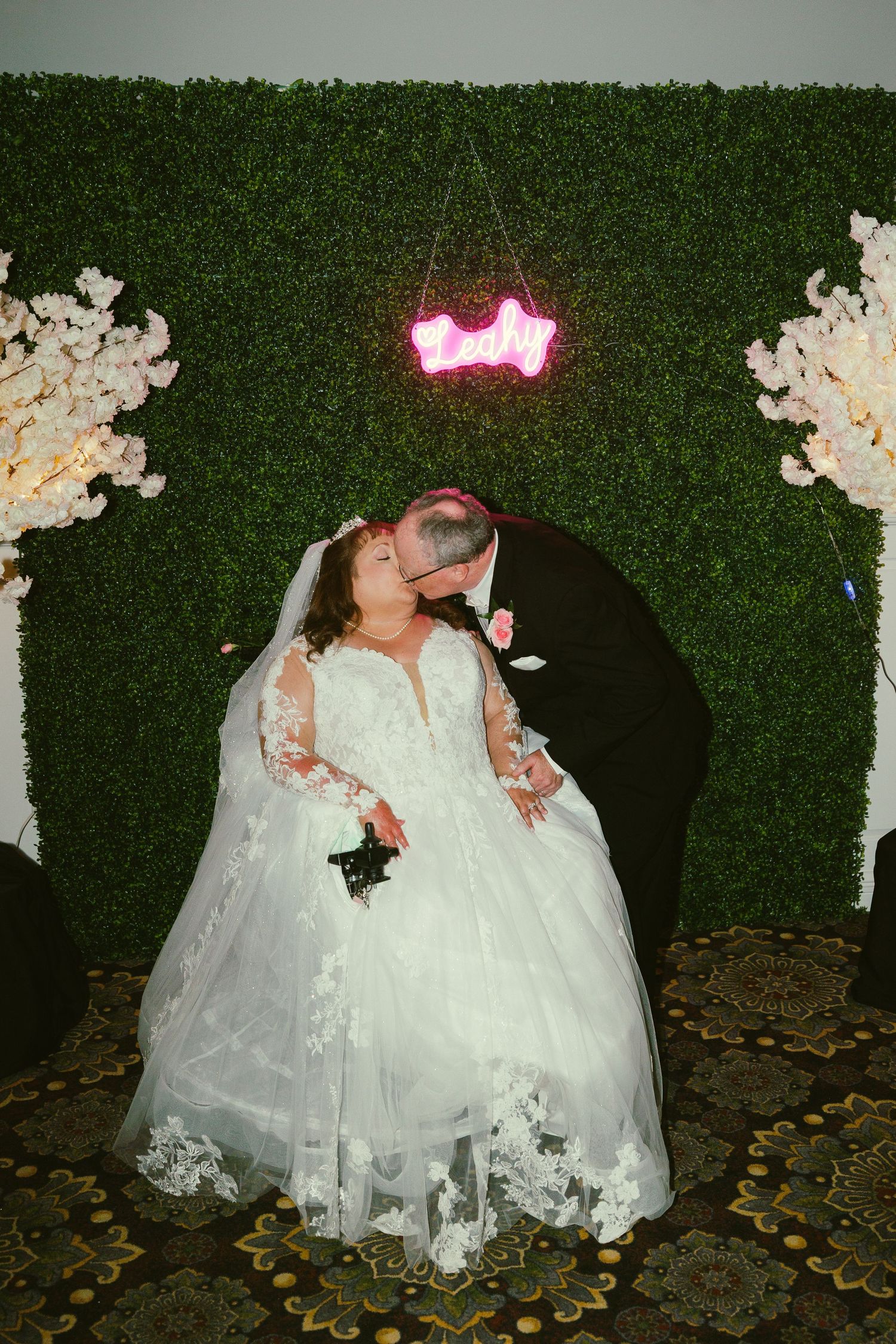 Wedding couple poses against a green hedge wall with decorative lights and floral arrangements.