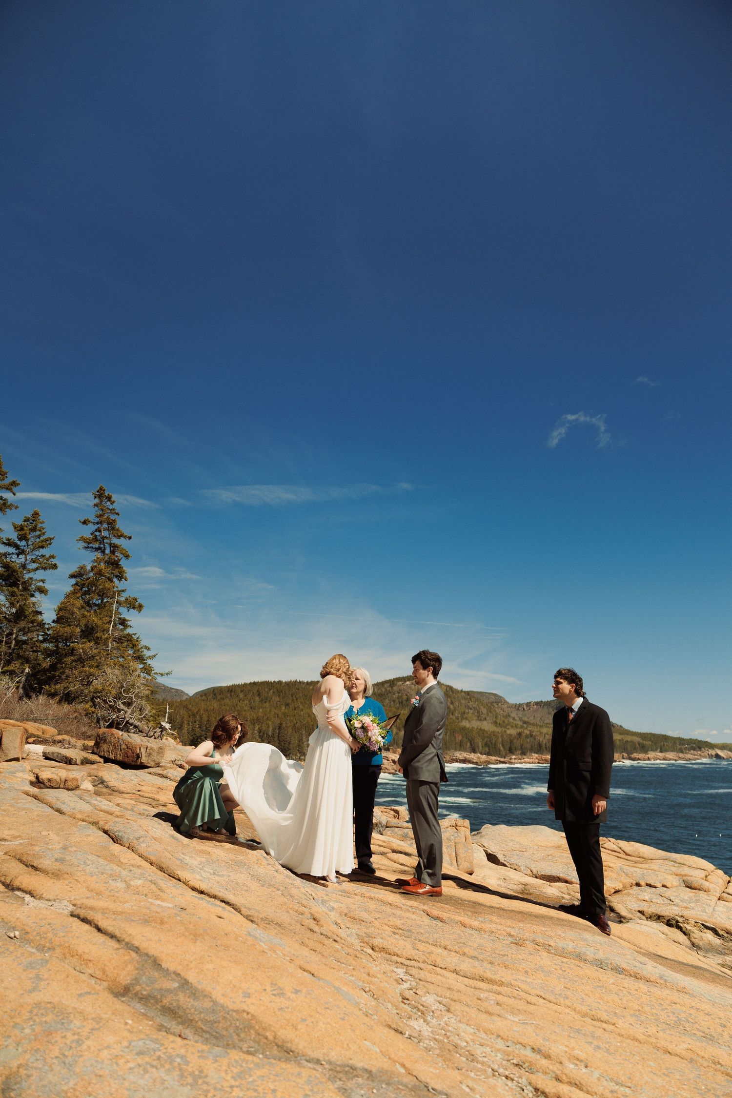Outdoor wedding ceremony on rocky coastal cliffs with ocean views, bride in white gown, groom, officiant, and guests under blue sky.