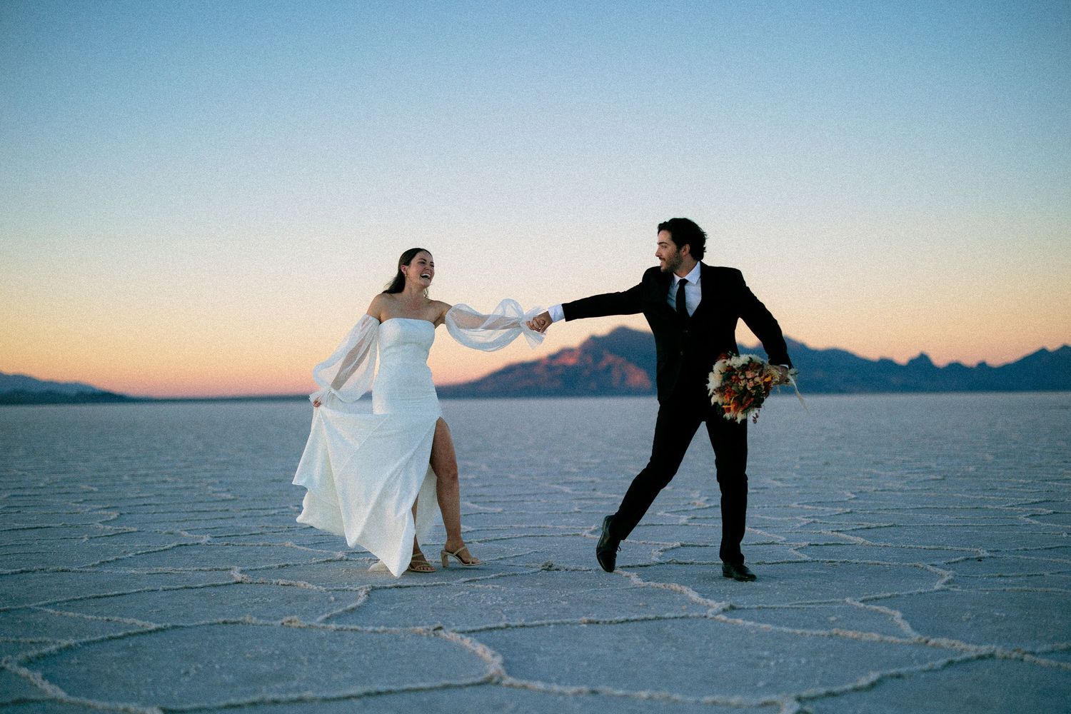 Playful wedding couple dances across white utah salt flats at sunset.