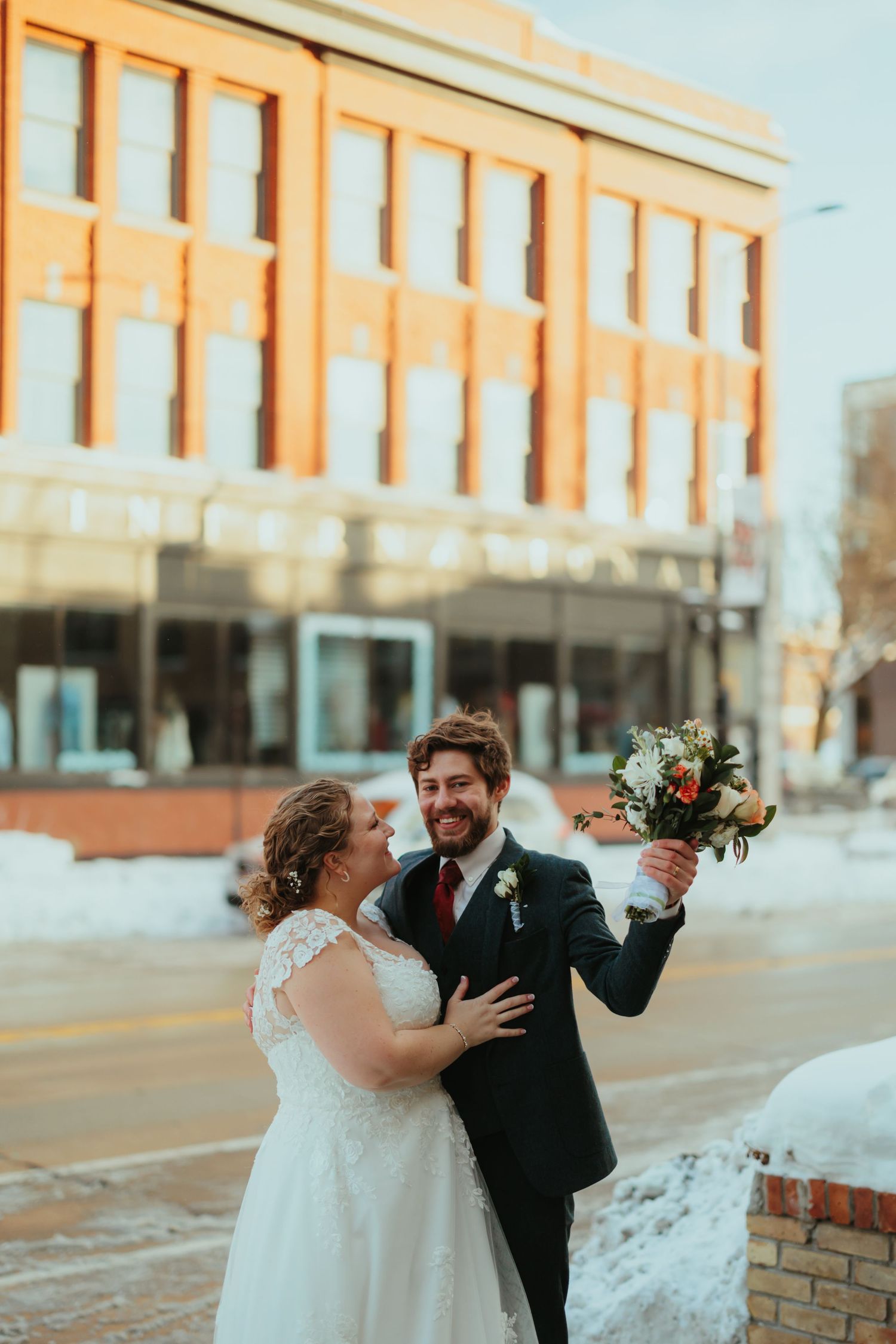 A joyful wedding couple shares candid moments together on a snowy winter day outside a brick building.