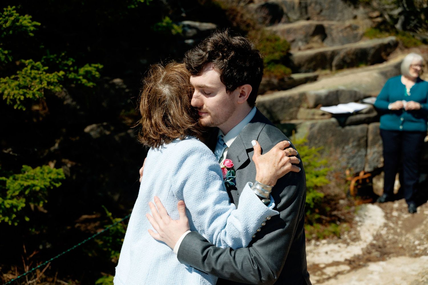 Couple embracing during outdoor wedding ceremony near rocky landscape with officiant in background.