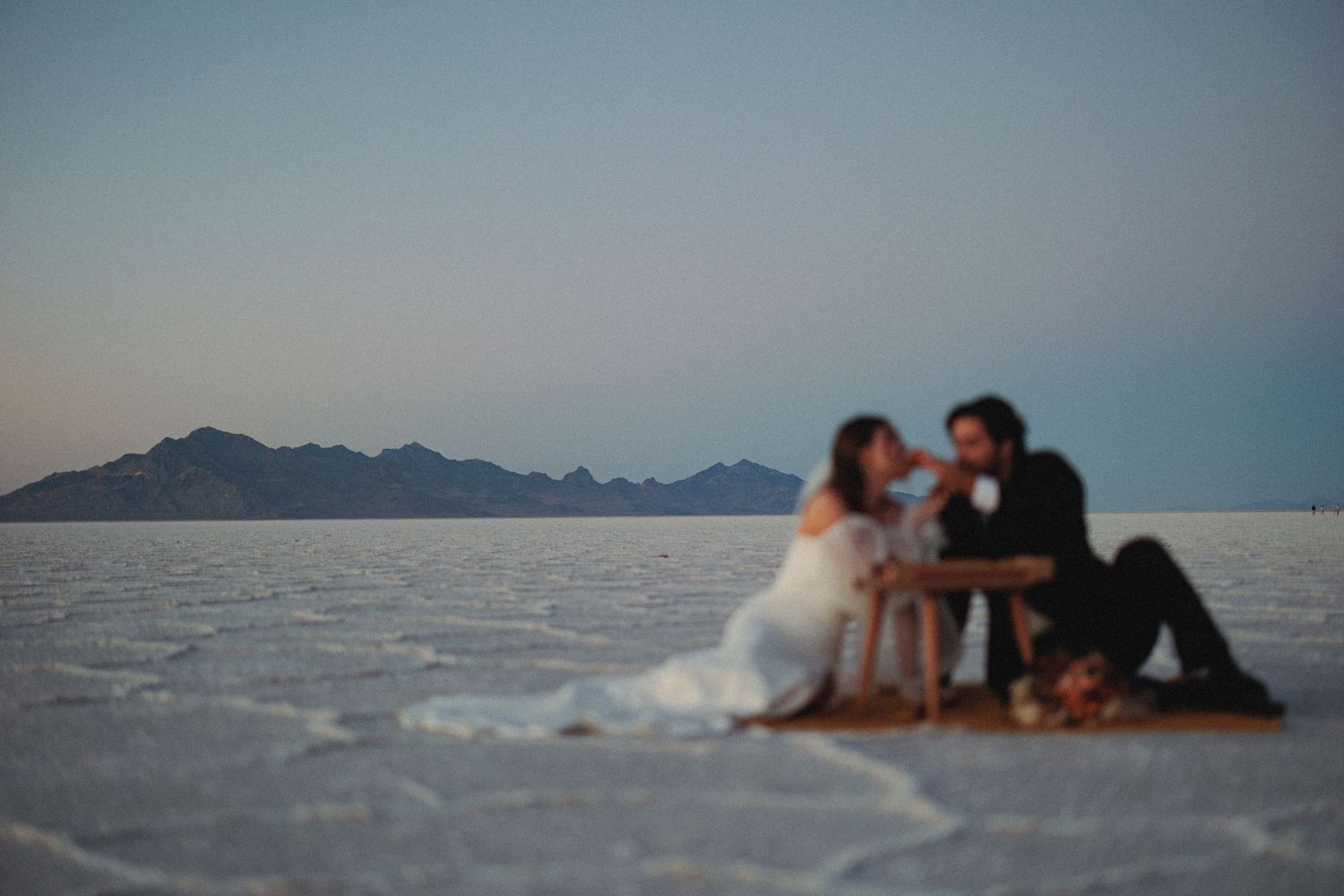 Wedding couple lounges on white utah salt flats with mountain silhouettes in the distance.