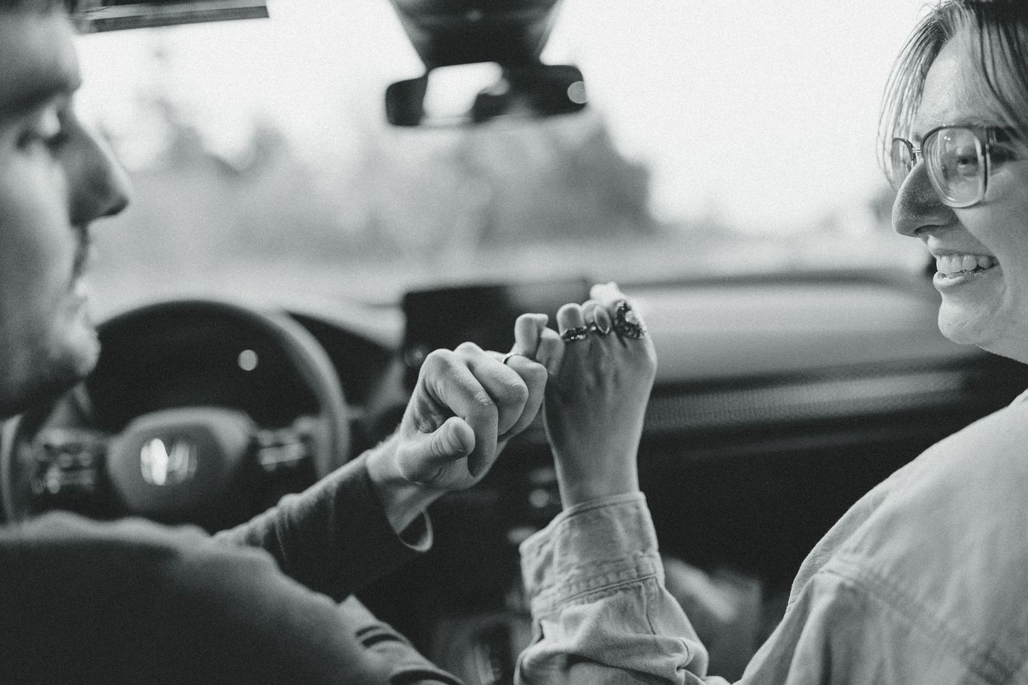 Black and white photo of a smiling couple holding hands inside a car, sharing a romantic moment.