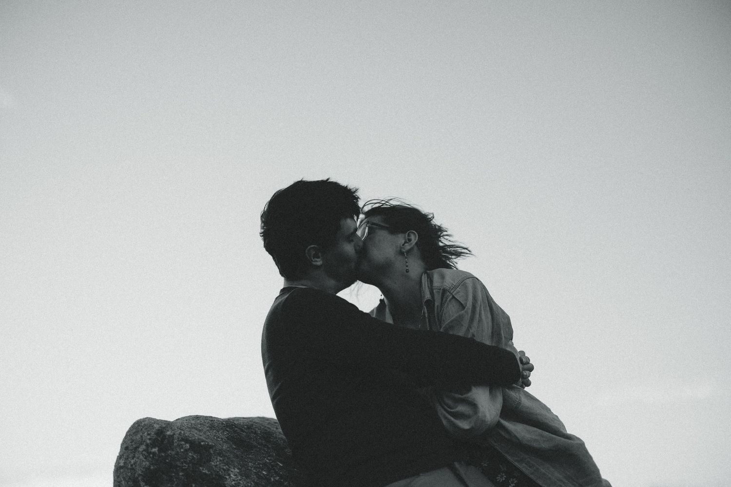 Black and white photo of a couple sharing a romantic kiss while sitting on rocks against a cloudy sky.