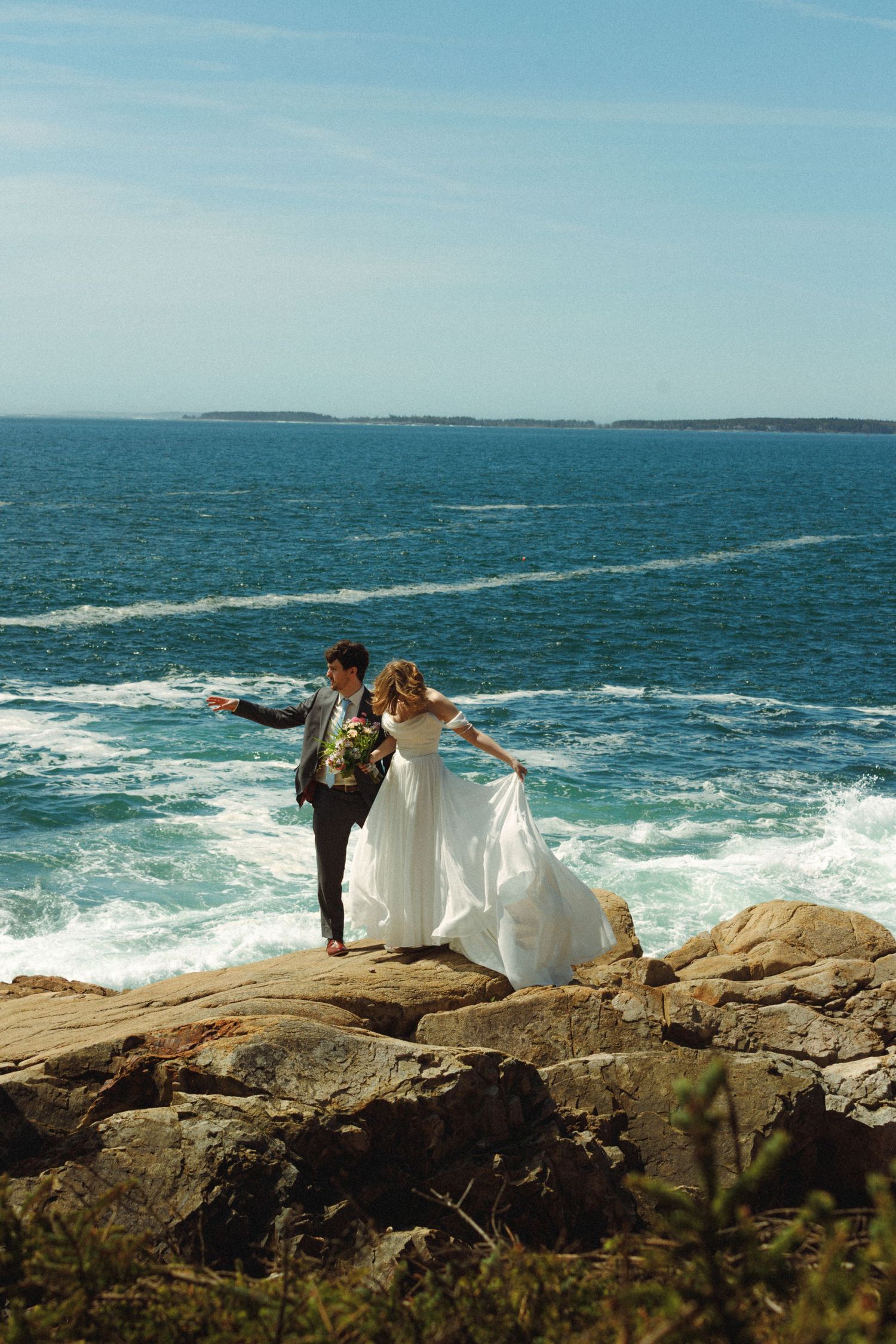 Bride and groom stand on rocky coastal cliffs with crashing ocean waves behind them on their wedding day.
