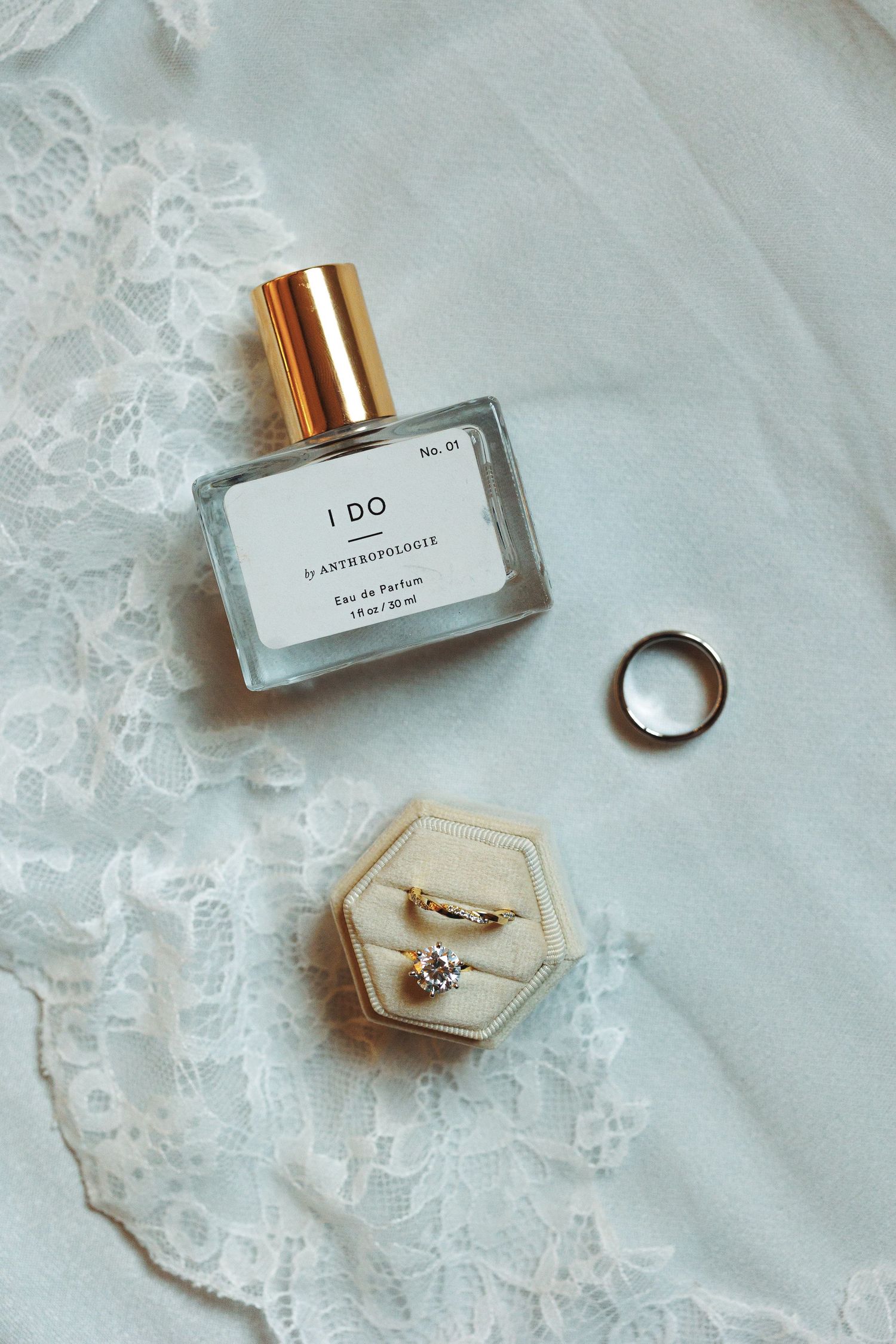 Wedding details including perfume bottle, rings and lace on white fabric.