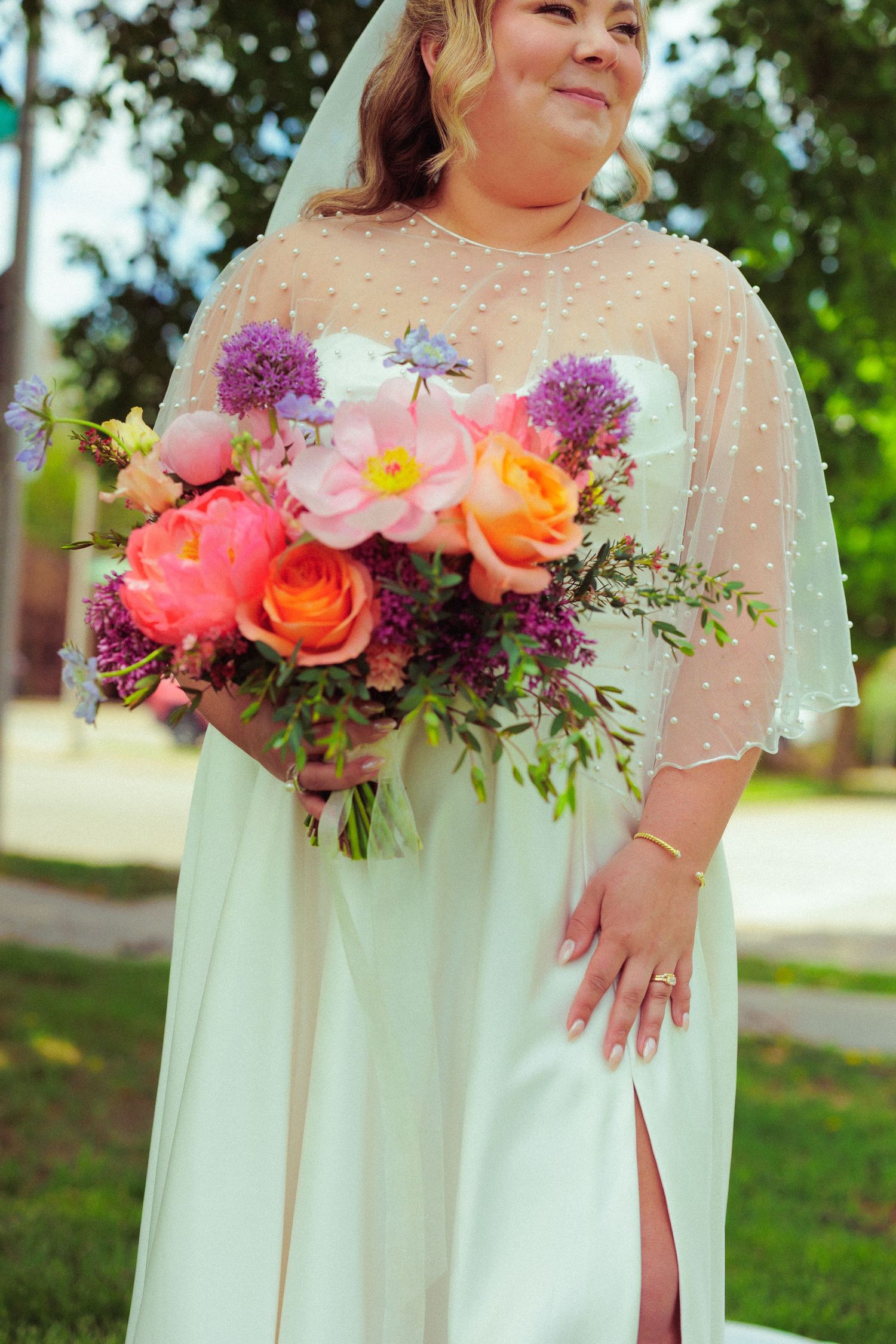 Bride in white dress with sheer sleeves holds vibrant pink and coral floral bouquet outdoors.
