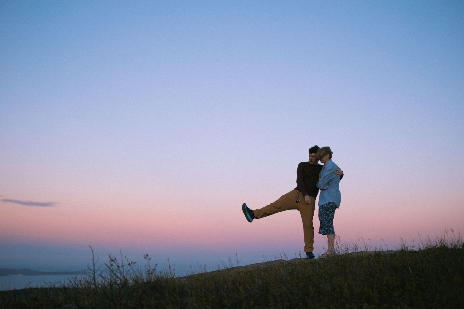 Romantic couple sharing a kiss on a hilltop at sunset with a colorful pink and blue sky backdrop.