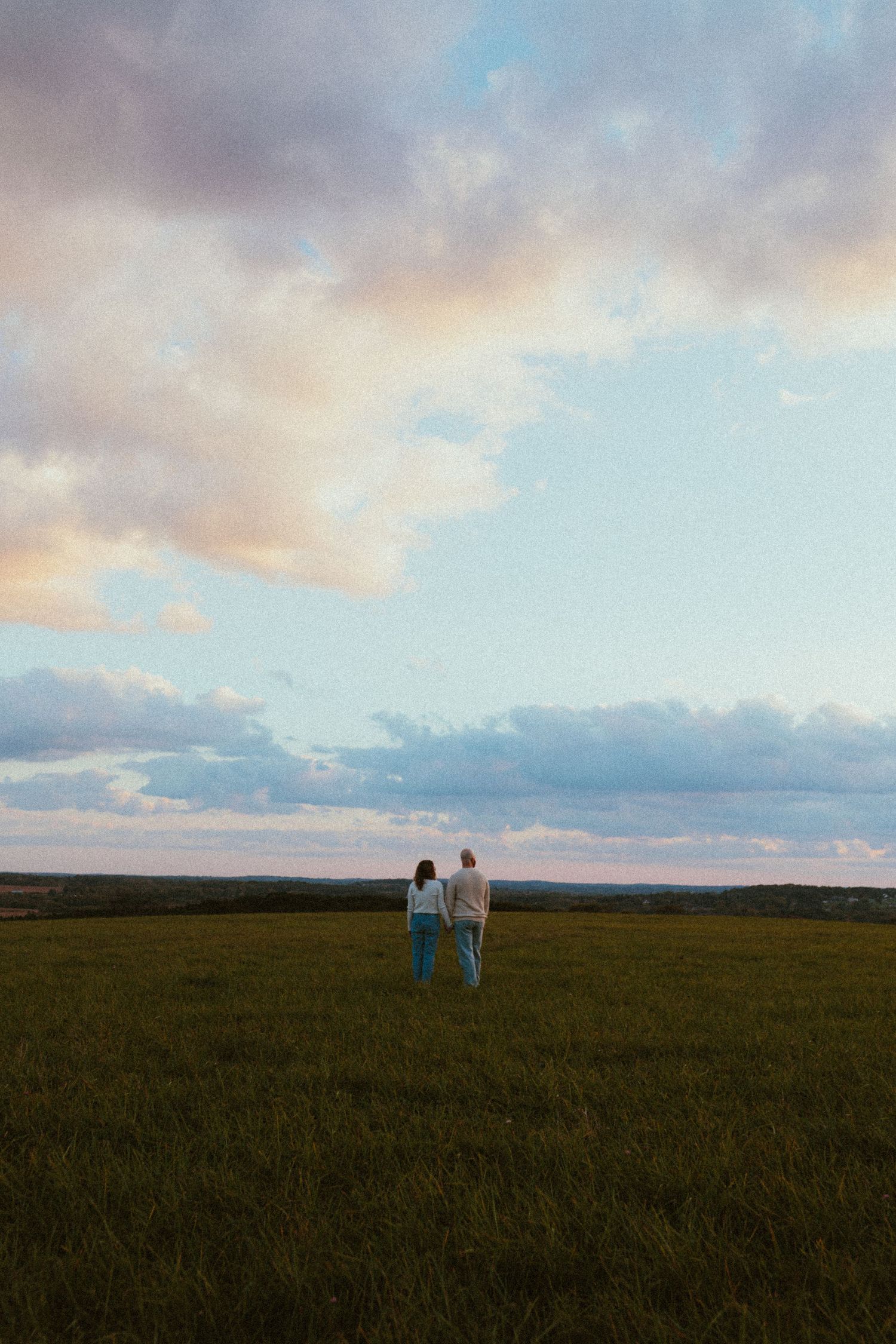 Couple walking together in a vast green field at sunset with dramatic clouds in the sky.