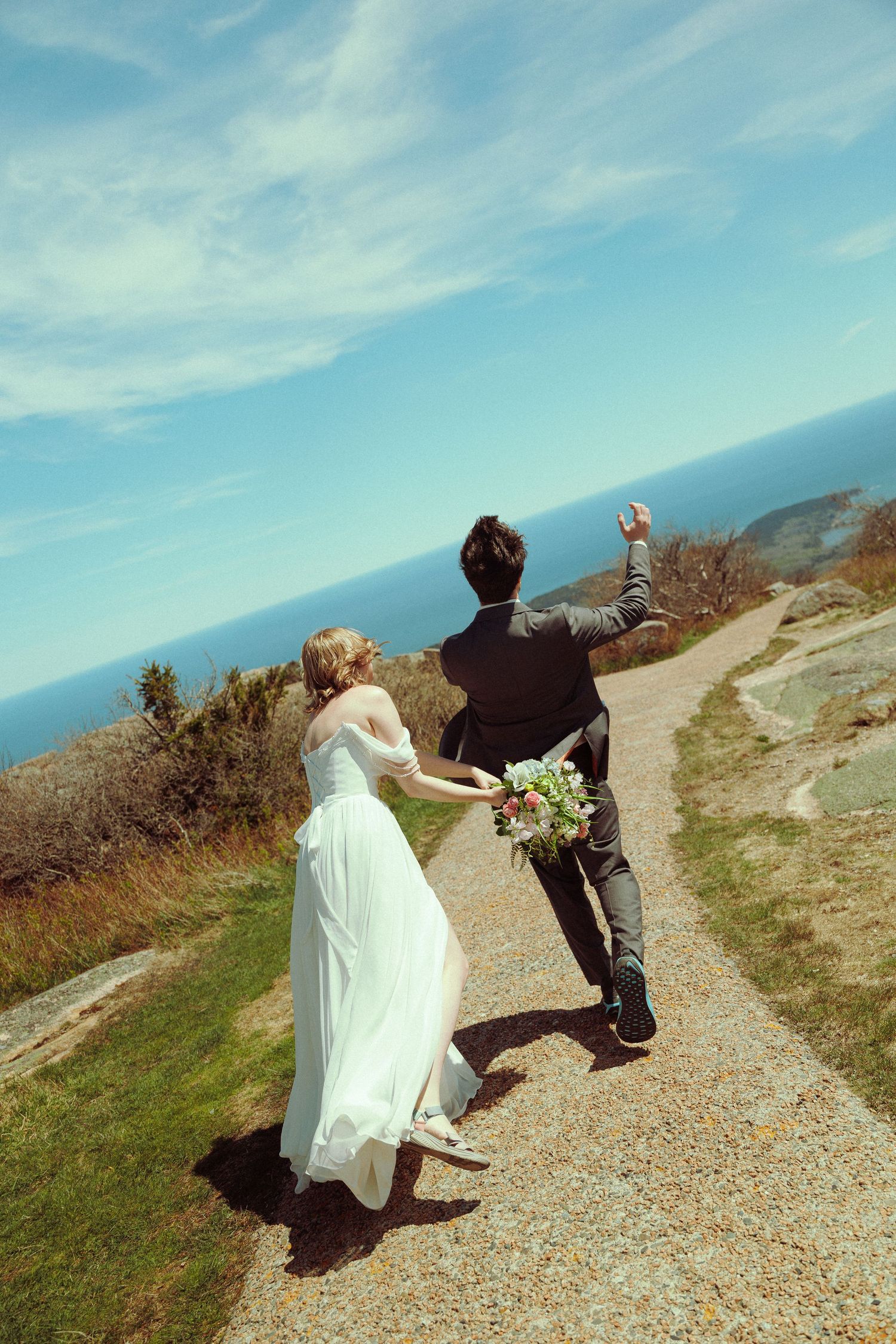Bride and groom walking hand-in-hand along a scenic coastal path, with the ocean horizon visible under a bright blue sky.
