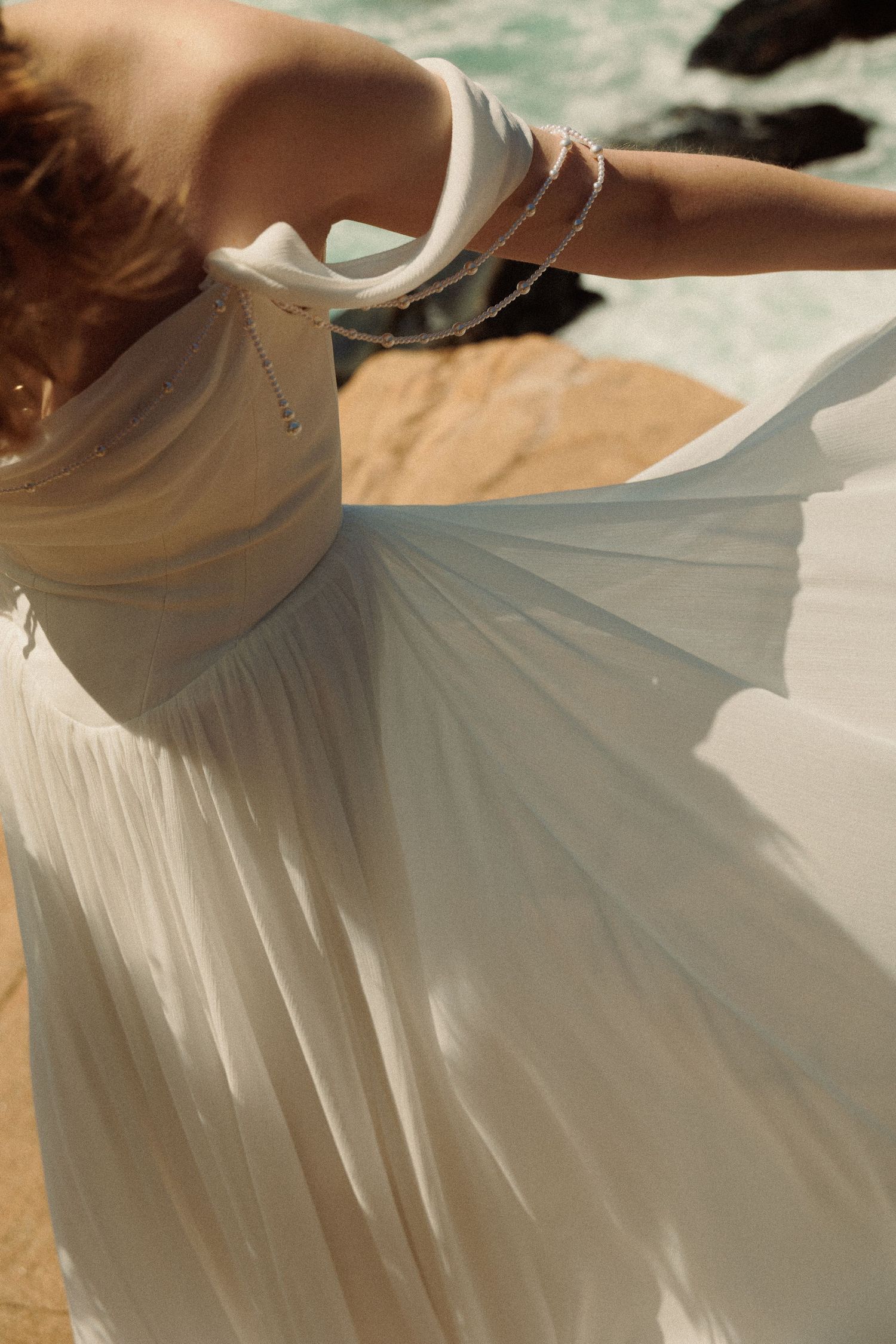 Bride in flowing white pleated wedding gown twirls on sandy beach with ocean rocks and waves in background.