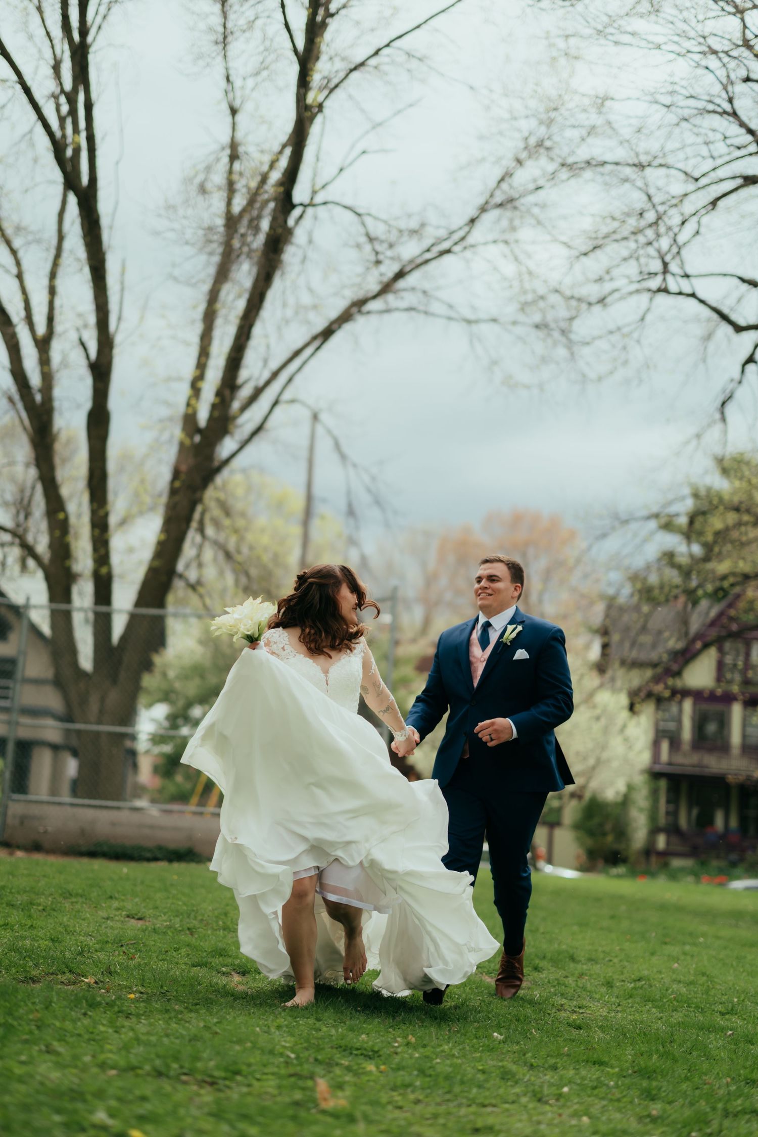 Playful wedding couple running and dancing on a grassy hill with bare trees in the background.