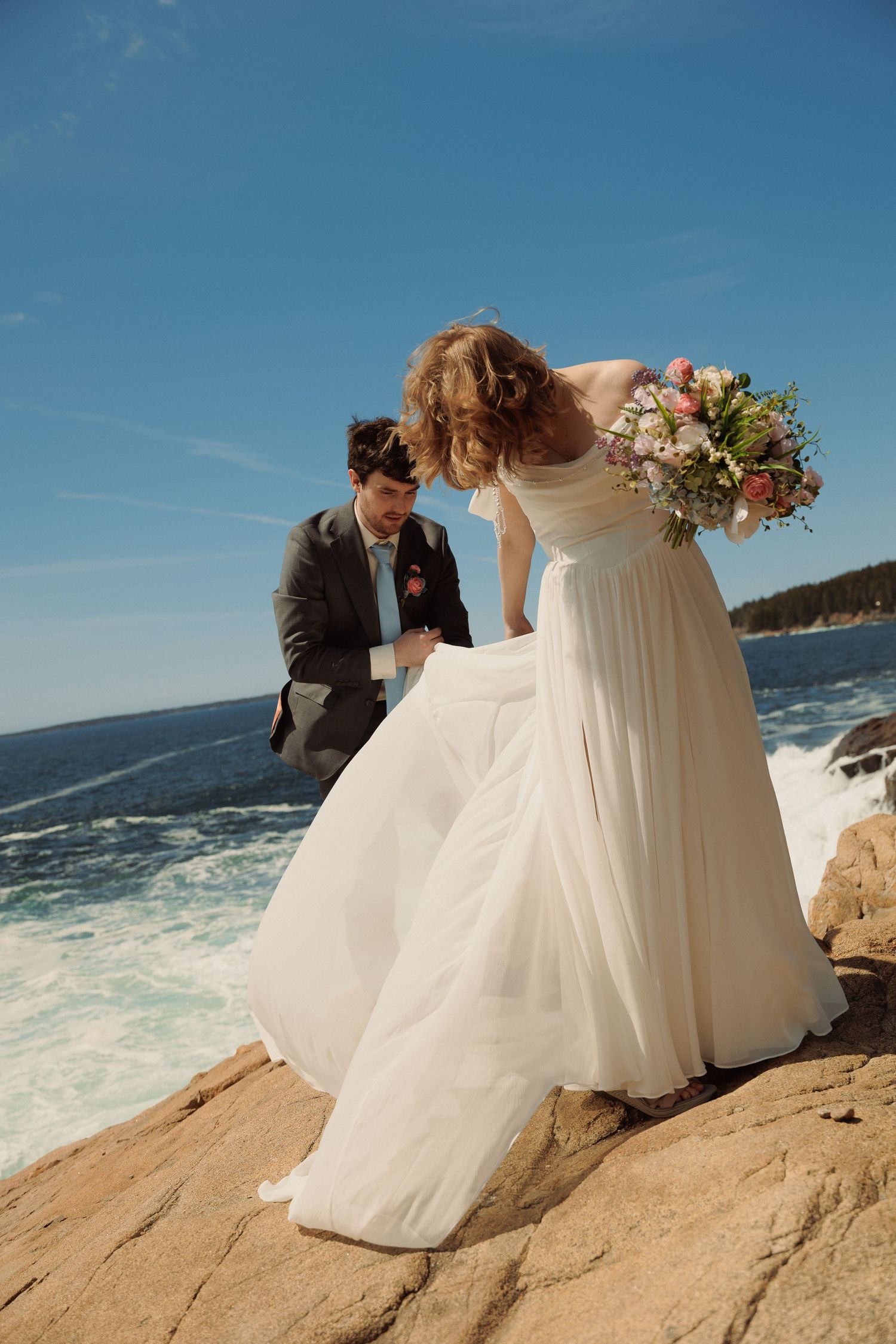 Bride in white gown holding bouquet with groom on rocky oceanside cliff under blue sky.