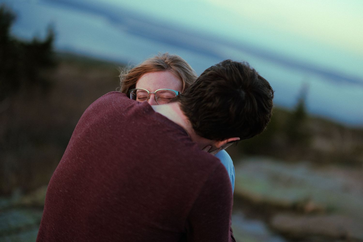 A woman with glasses embracing a man in a maroon sweater outdoors near water at dusk.