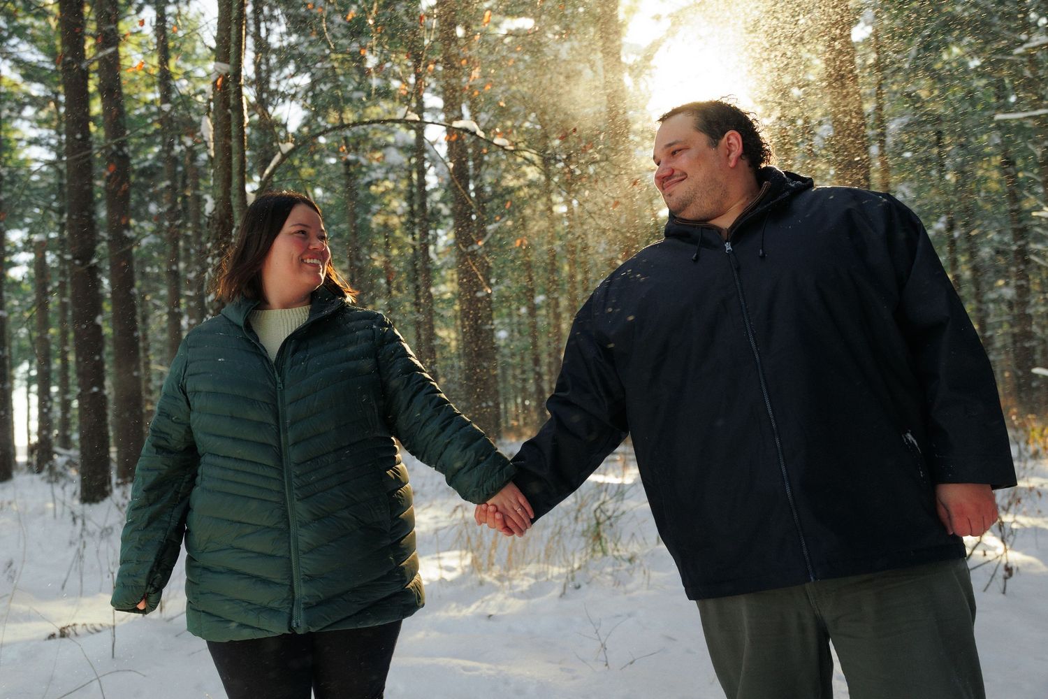 Couple walking through a snowy forest path with sunlight filtering through the trees.