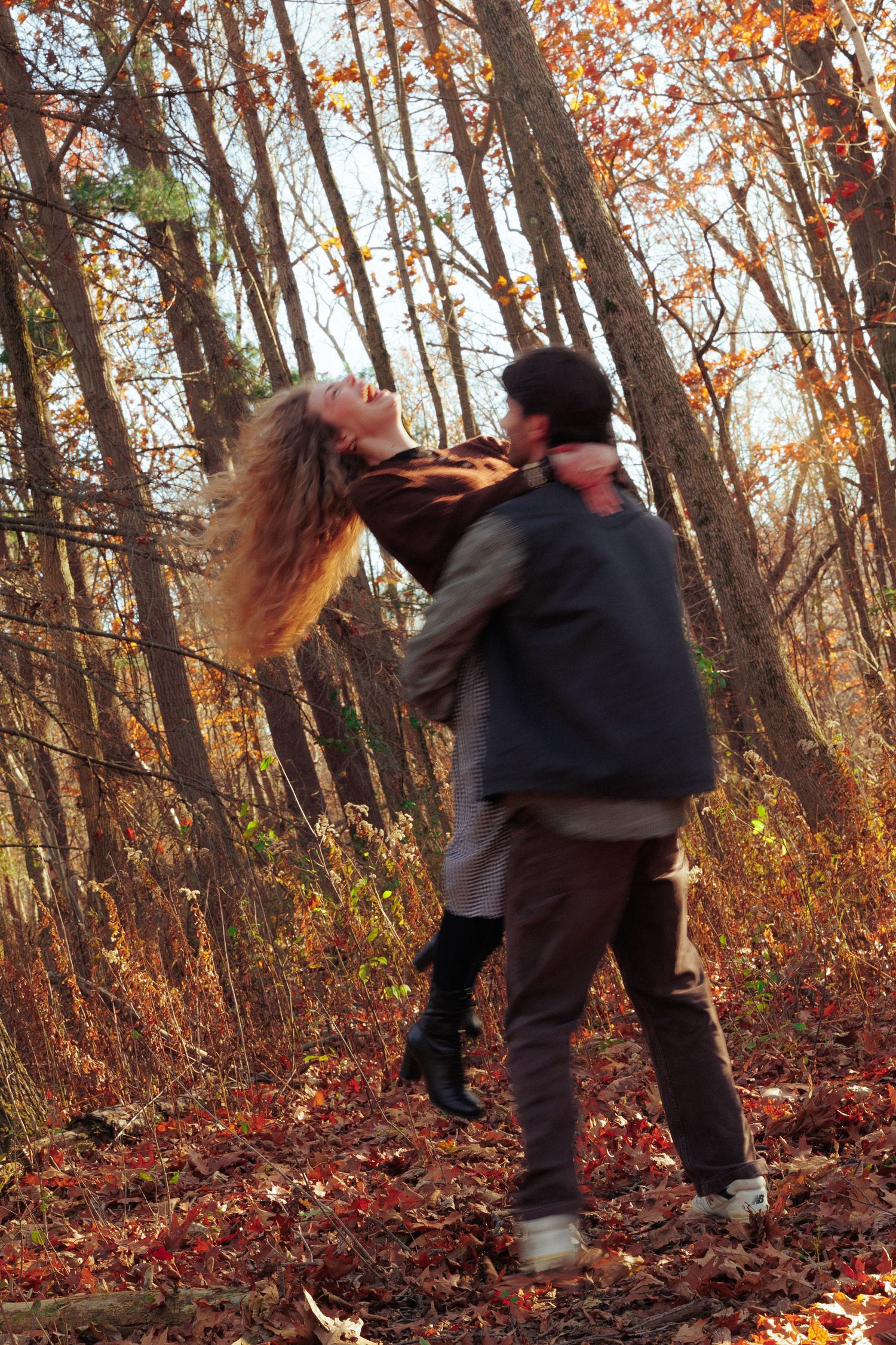 A couple playfully interacting among autumn trees with fallen orange leaves in a forest setting.