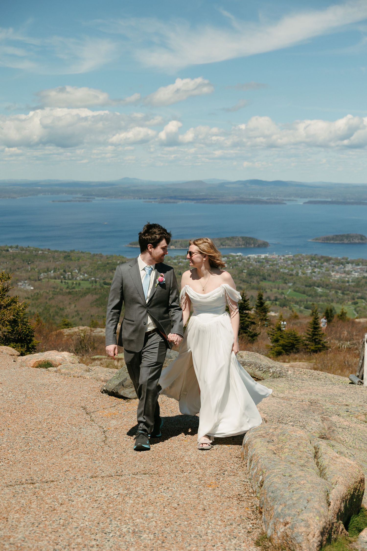 Bride and groom hold hands walking on a mountain summit with panoramic water views behind them.