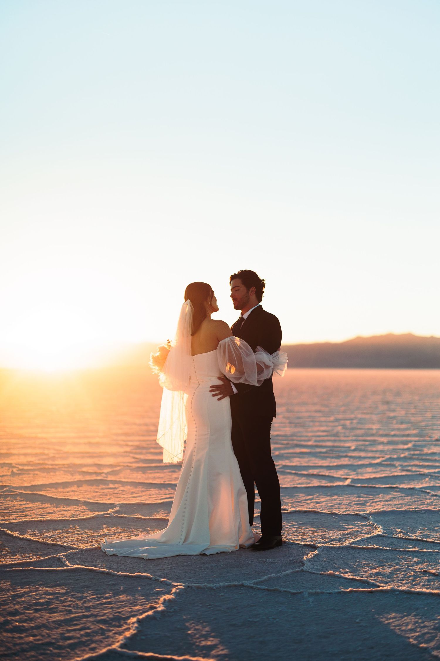 Wedding couple poses on crystalline utah salt flats during golden hour with mountains in background.