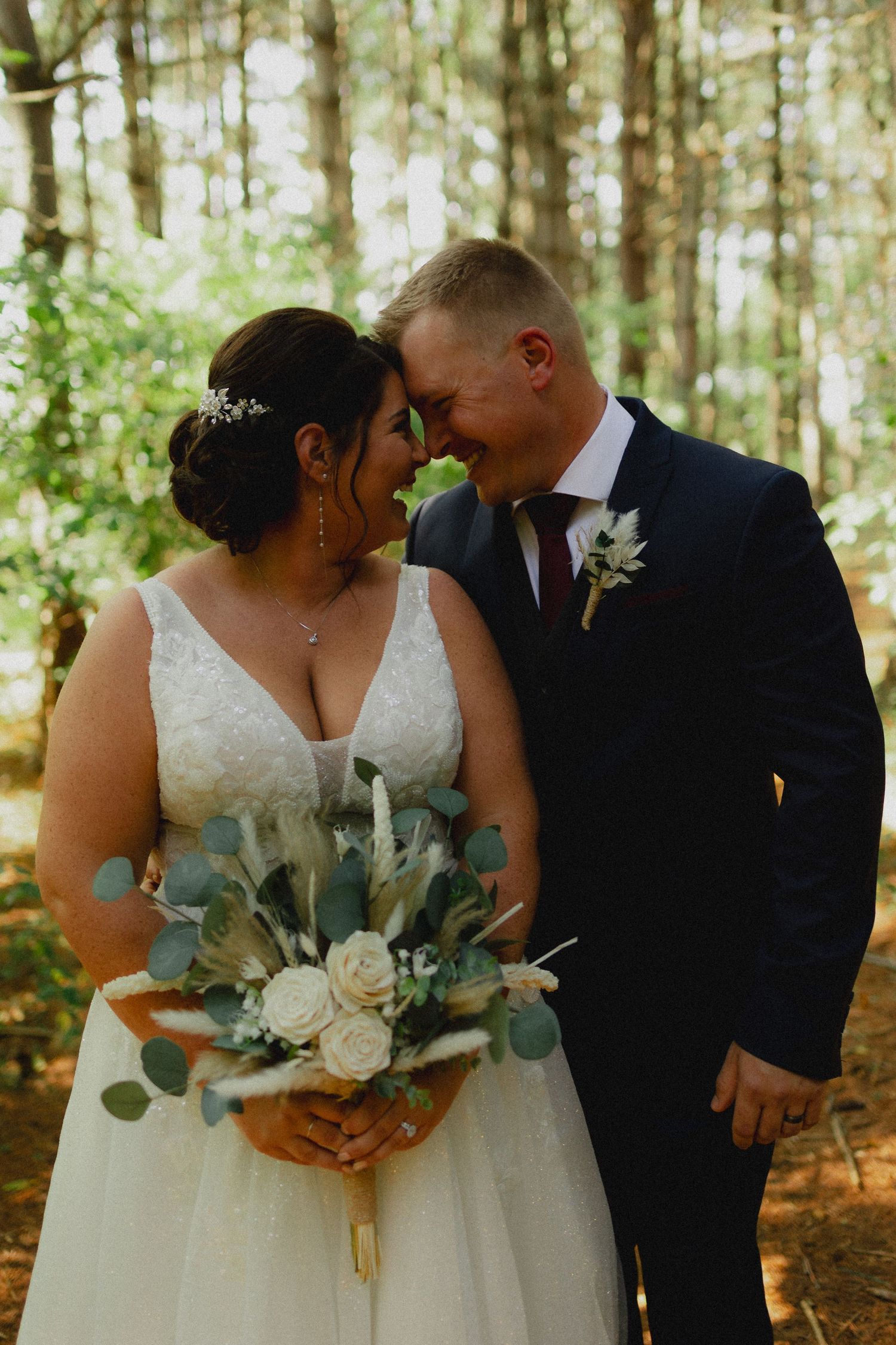 A romantic wedding couple shares intimate moments in a sunlit forest while holding a white and green floral bouquet.