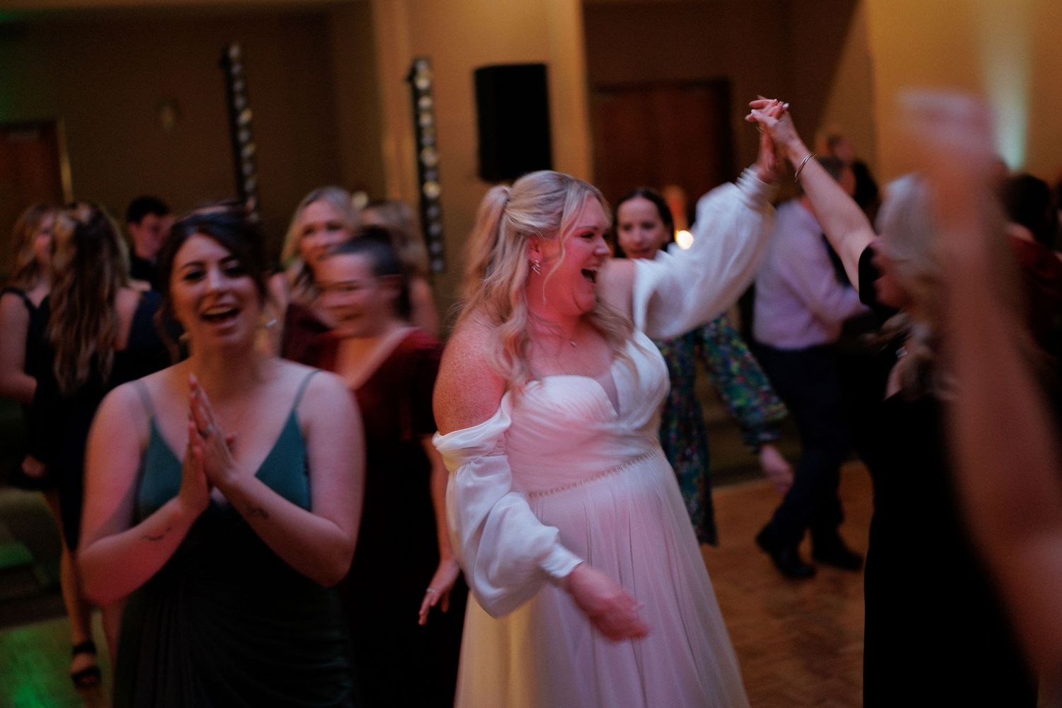 Wedding reception guests dancing in a dimly lit venue with warm lighting.