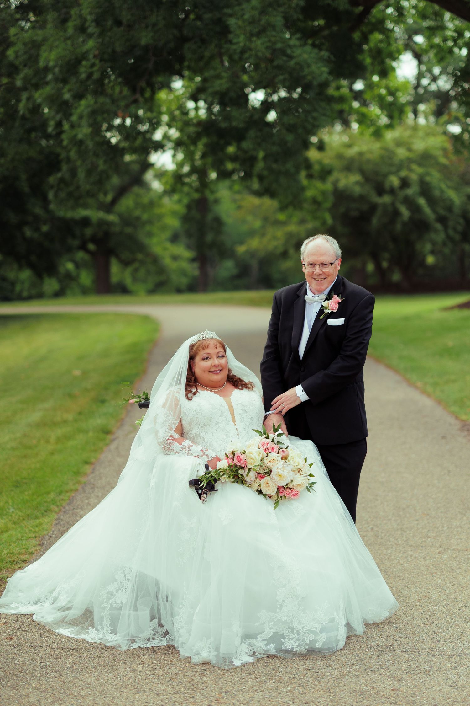 Bride in flowing lace gown poses with partner on garden pathway.