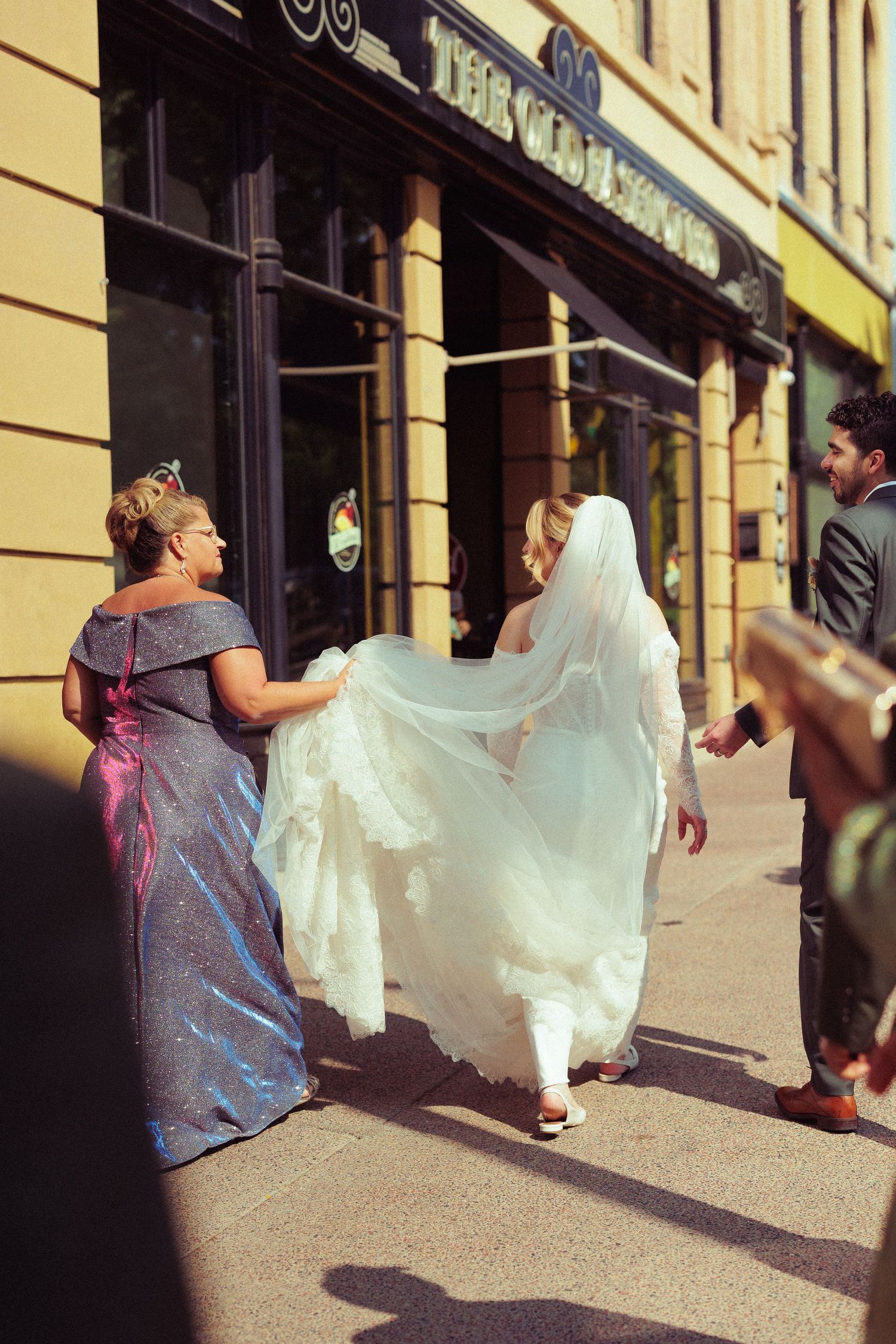 Wedding dress train flowing behind as people walk along a sunlit urban sidewalk.