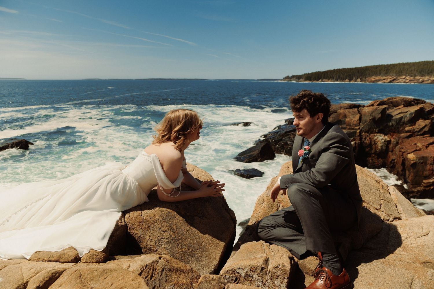 Bride and groom sit on rocky coastal cliffs overlooking ocean waves on their wedding day.