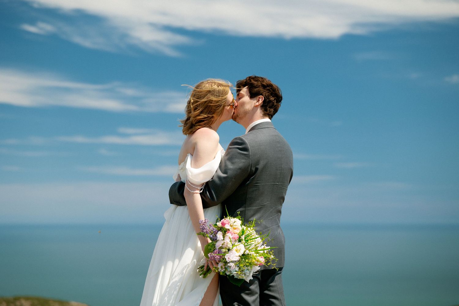 Bride and groom sharing a romantic kiss outdoors with ocean and blue sky backdrop on their wedding day.