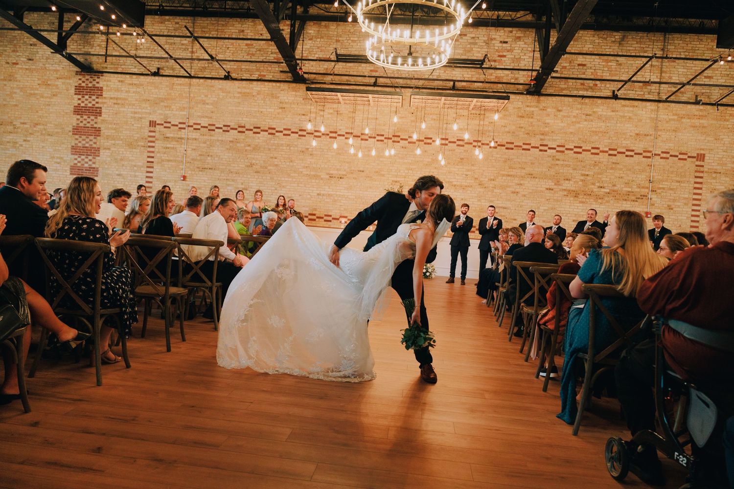 Newlyweds share their first dance in a rustic venue with string lights and exposed brick walls.