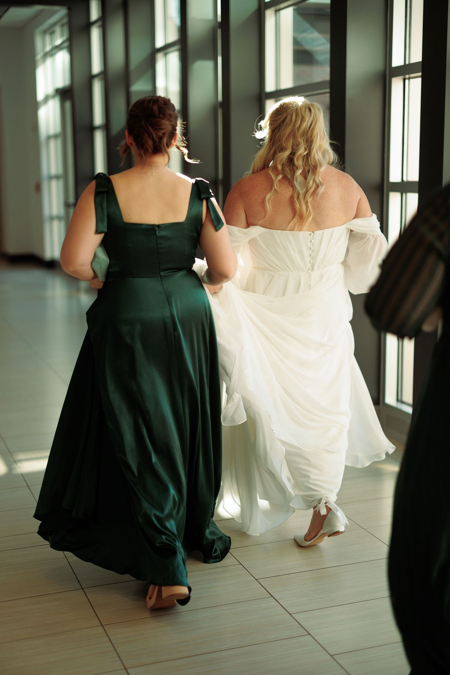 Bride in white dress and bridesmaid in dark green dress walking down a sunlit hallway.