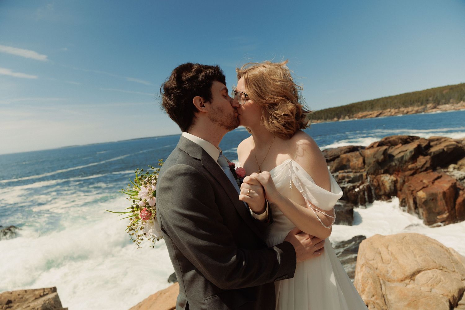 A bride and groom share a romantic kiss on rocky coastal cliffs with crashing ocean waves under a bright blue sky.