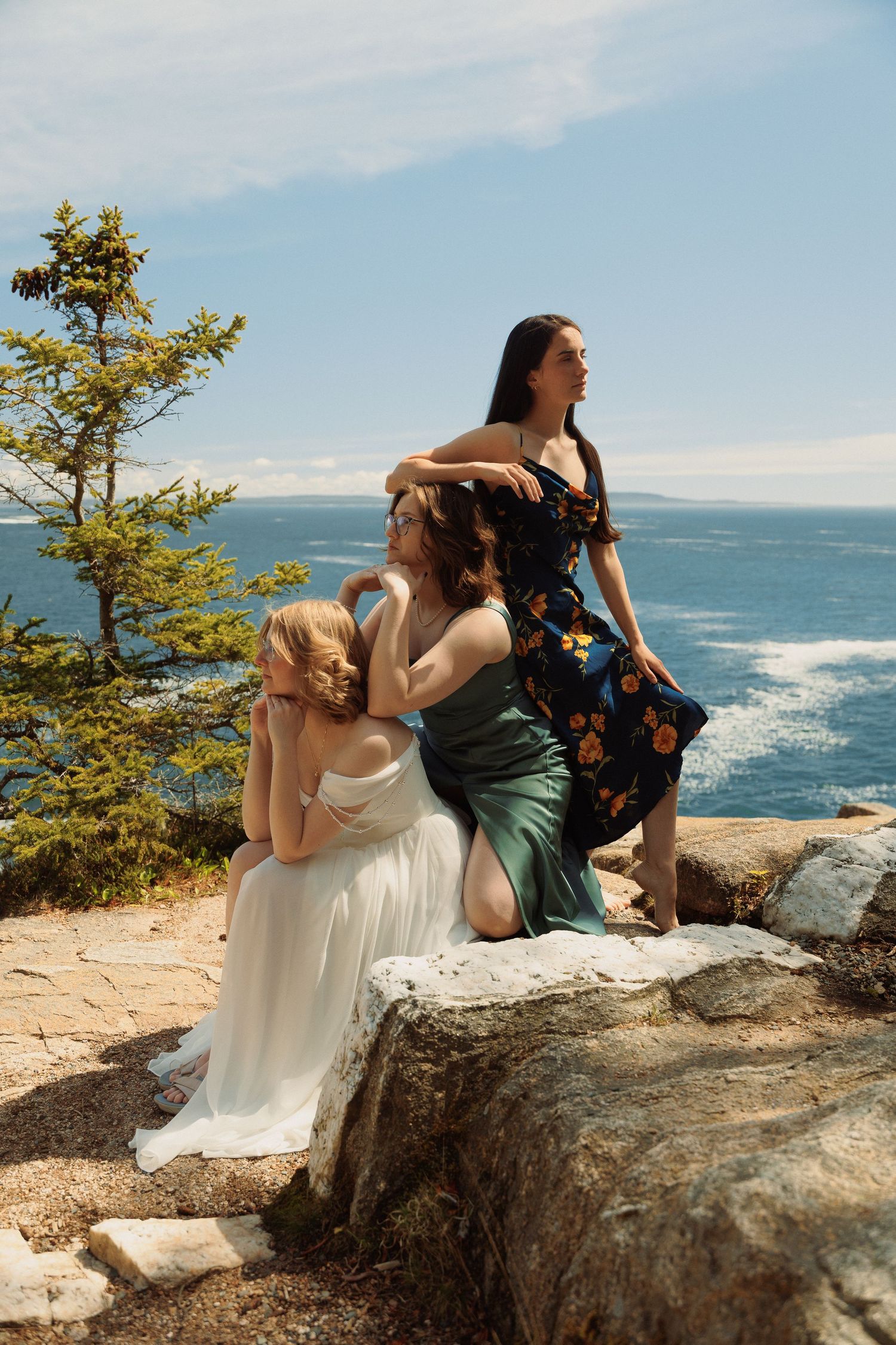 Three women in elegant dresses pose on coastal rocks with ocean views behind them on a sunny day.