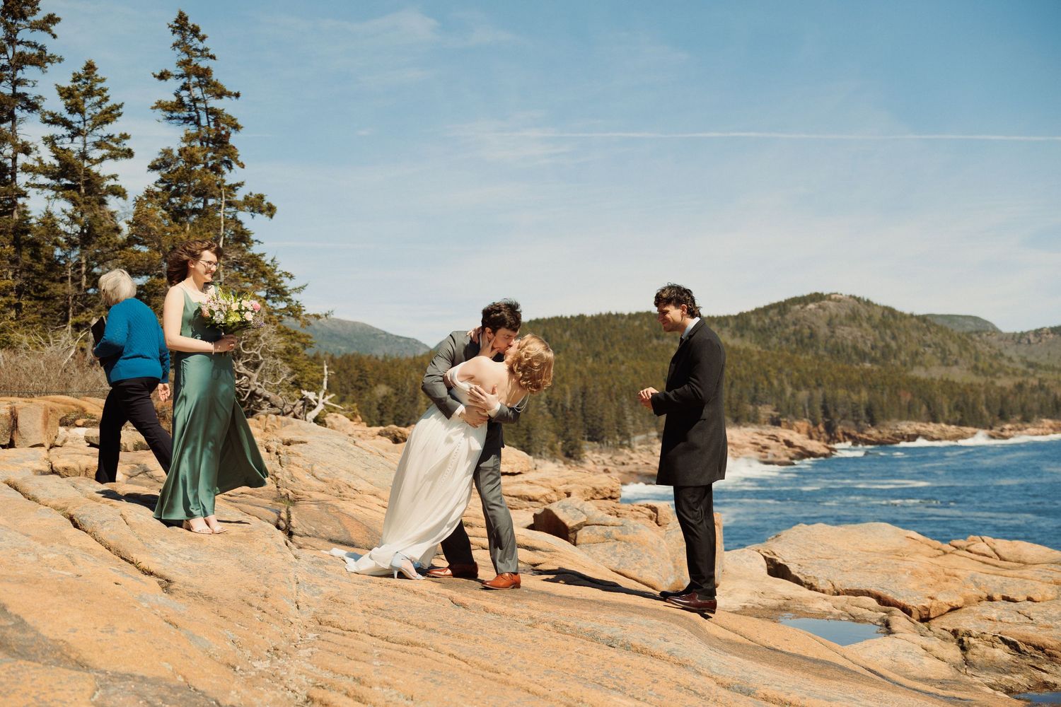 Couple shares first kiss during outdoor wedding ceremony on rocky Maine coastline with bridesmaid and officiant nearby.