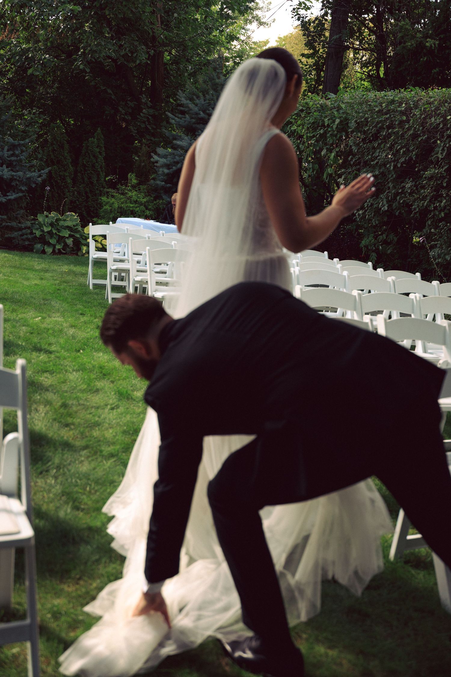 Couple in formal wedding attire playfully posing with white chairs outdoors.