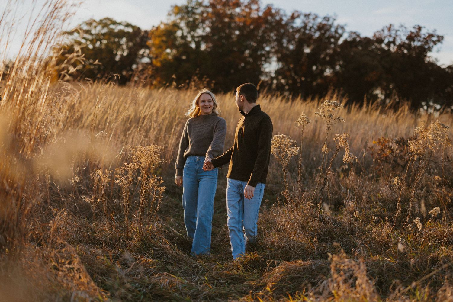 Couple walking through tall grass during golden hour with warm autumn lighting.