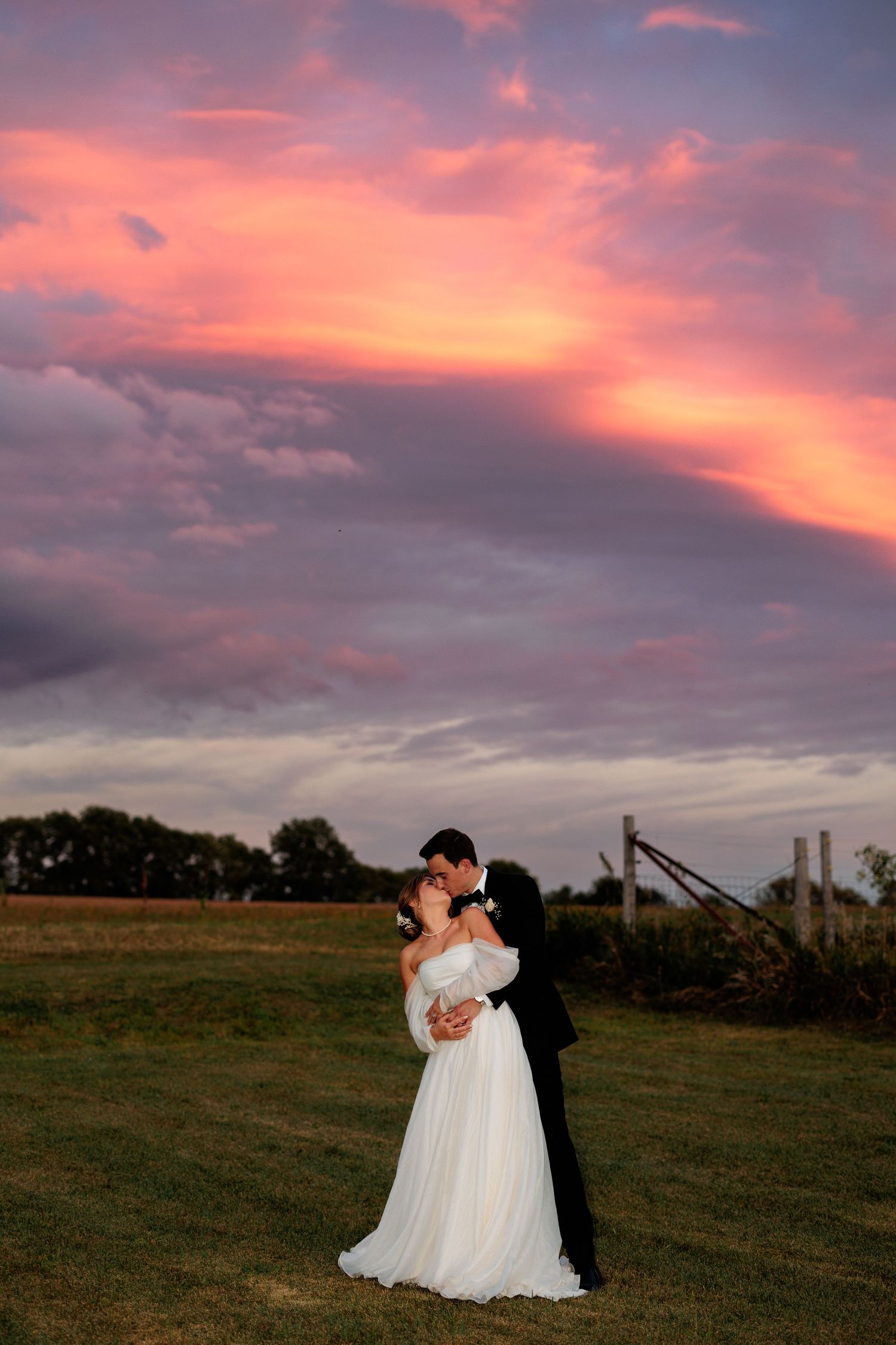A couple shares an intimate moment during sunset in an open field with dramatic pink and orange sky.