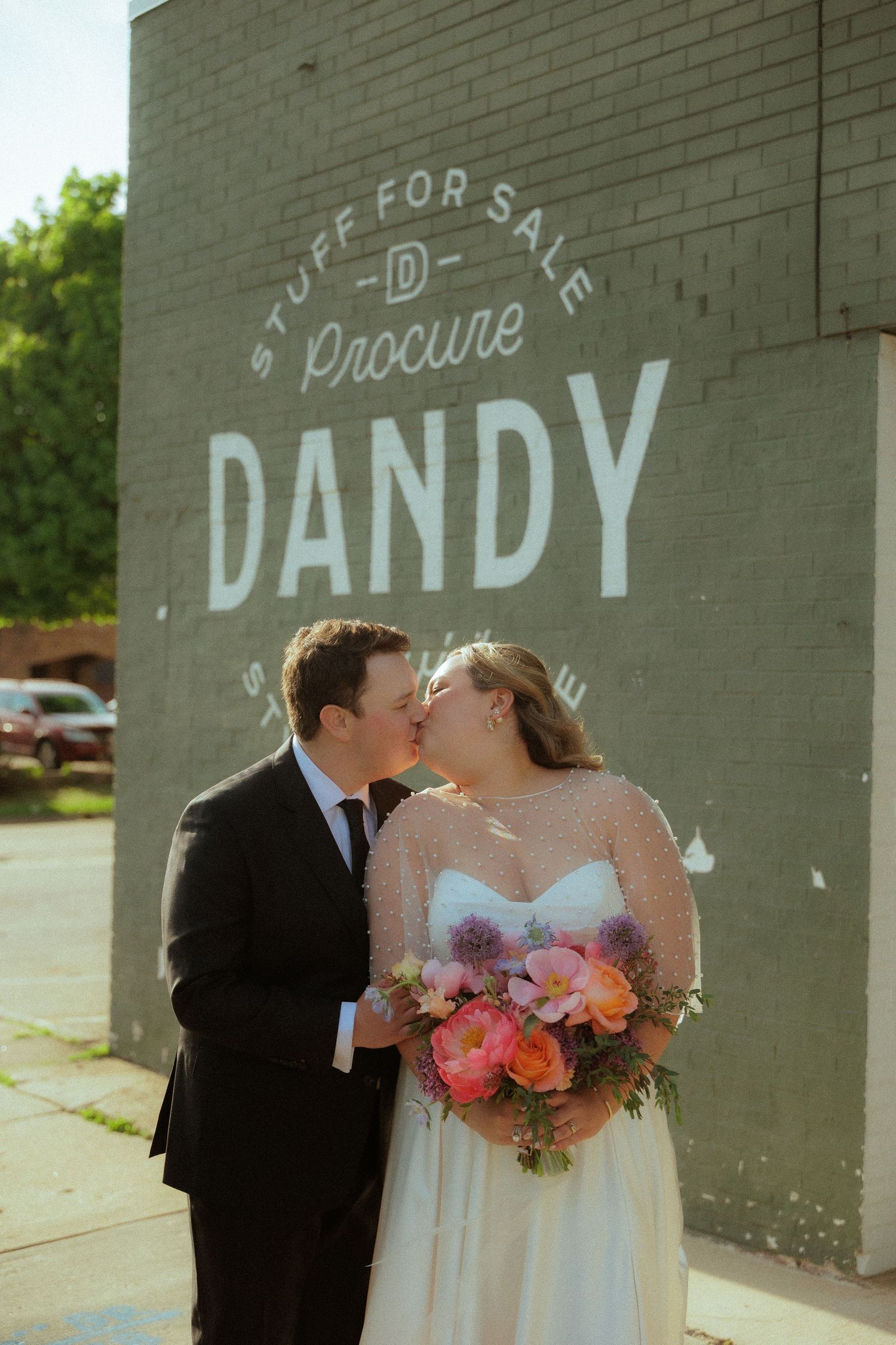 Newlyweds pose for photos against grey brick wall with 'DANDY' text.