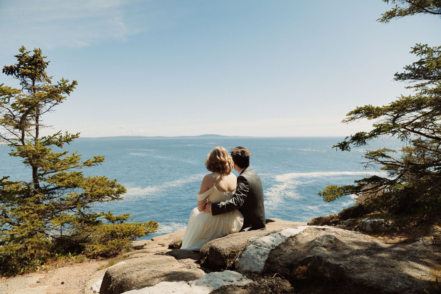 Bride and groom sitting on rocky cliff overlooking blue ocean, surrounded by pine trees on a sunny wedding day.