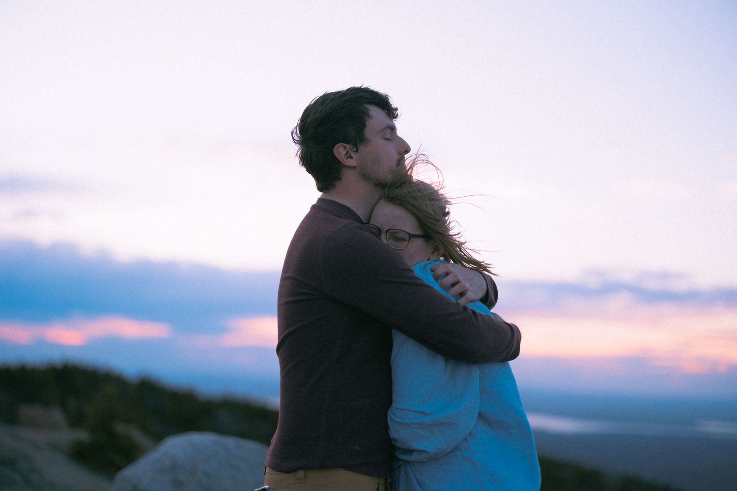 Couple embracing at sunset on a mountain overlook, sharing a romantic moment against a colorful twilight sky.