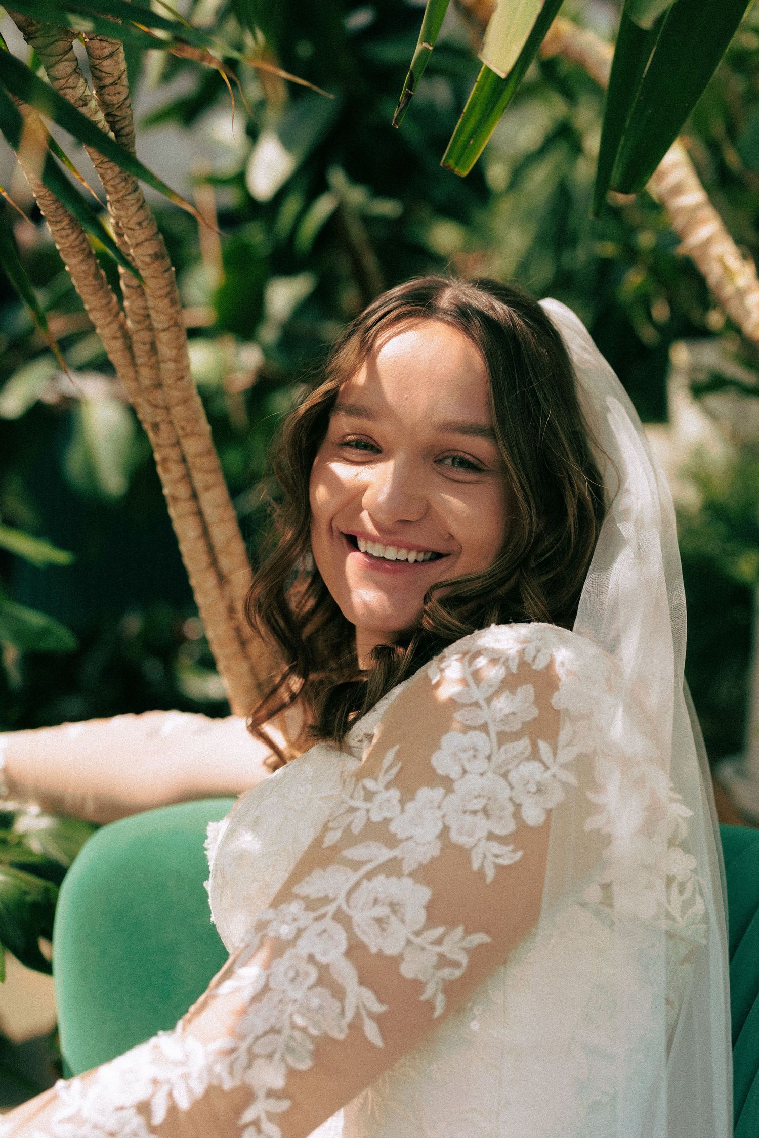 A bride in a lace wedding dress poses outdoors among leafy trees.