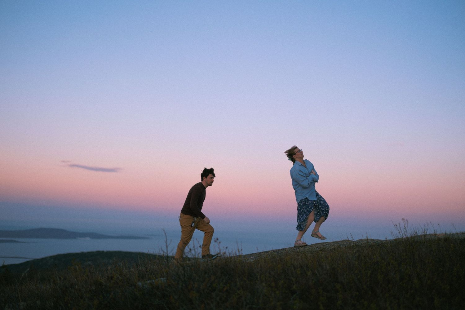Two people dancing on a grassy hill at dusk with a pink and blue sunset sky over the ocean.