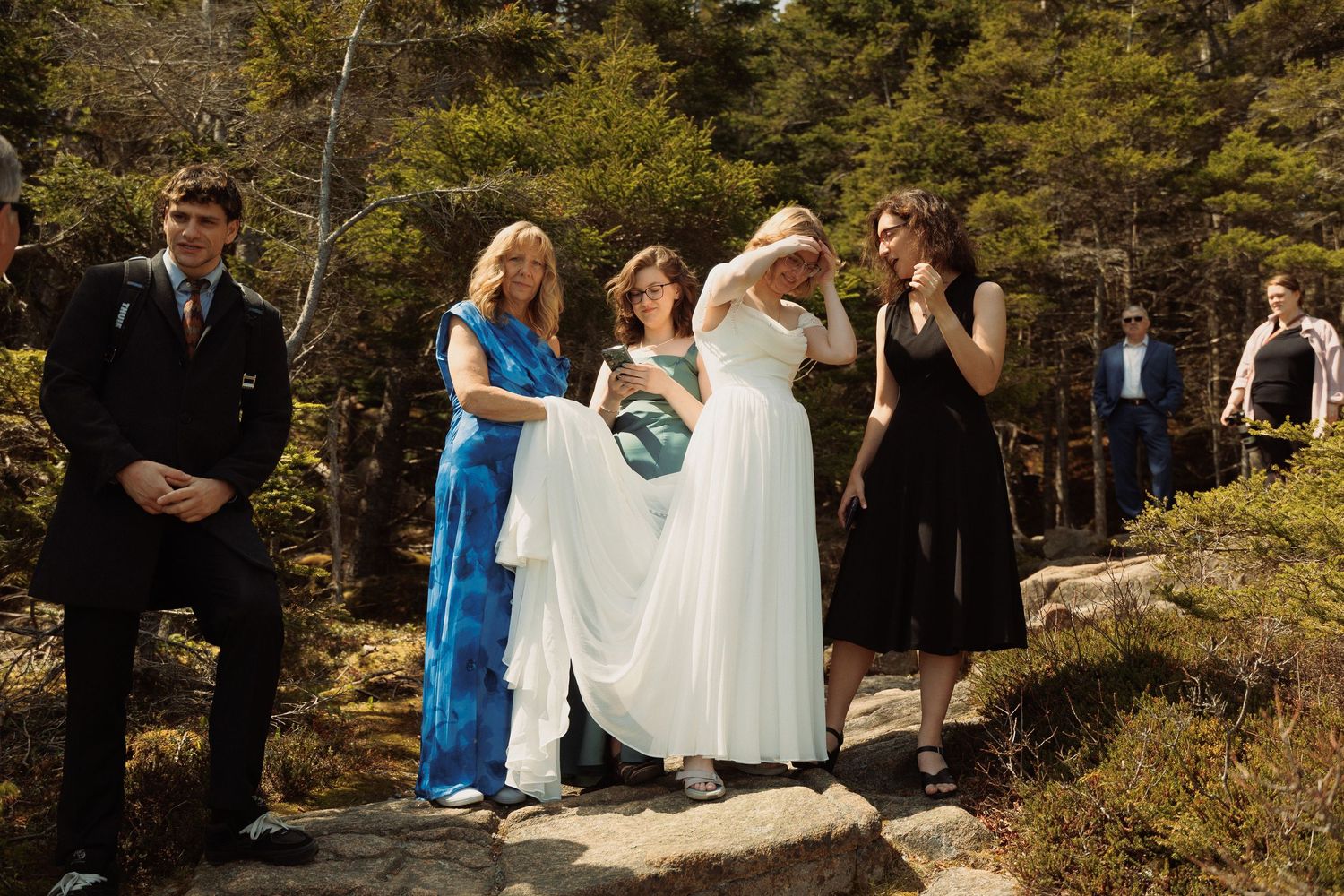 A bride in white gown stands with family and friends on rocky outdoor terrain surrounded by trees at a wedding ceremony.