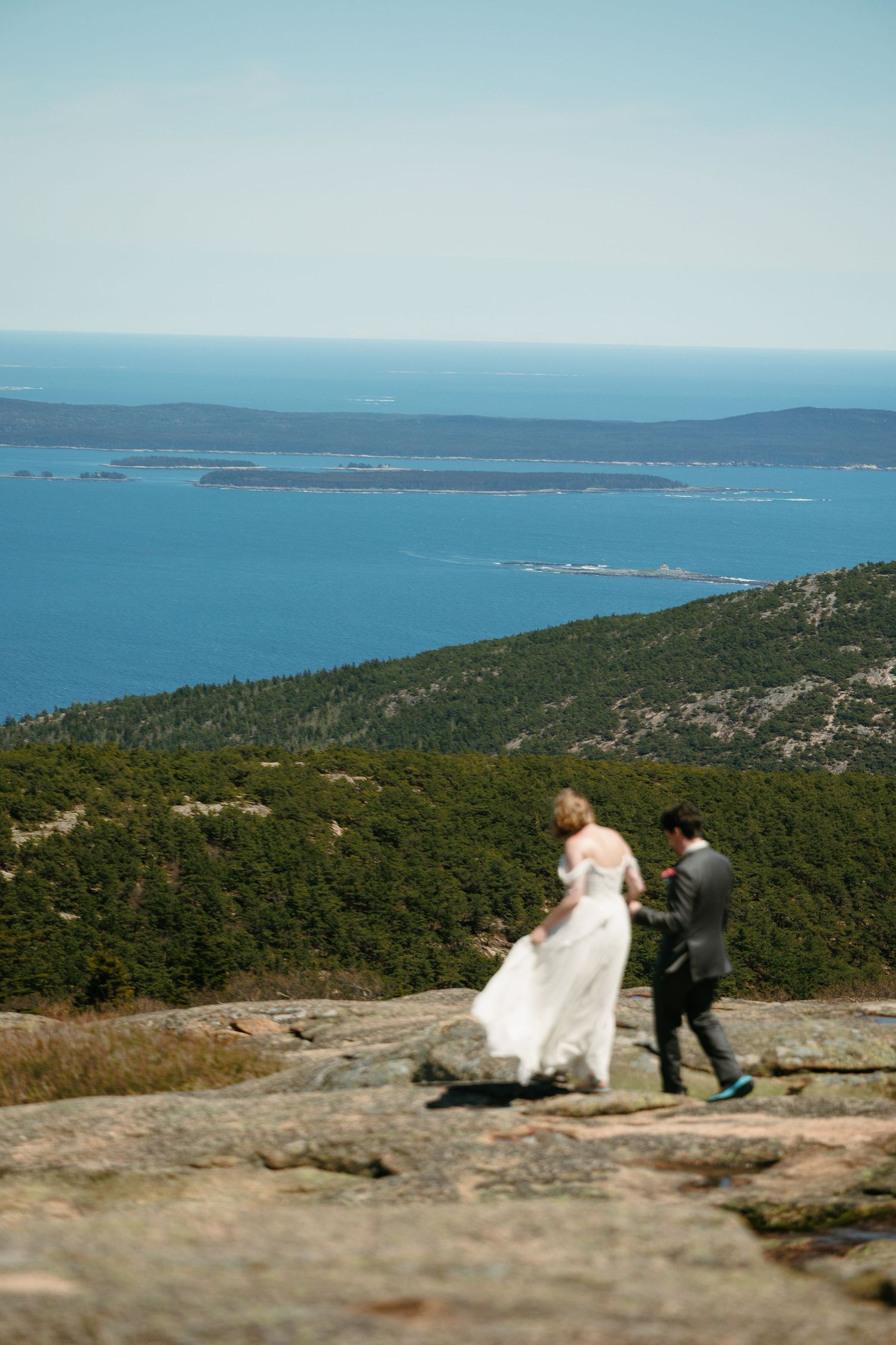 Bride and groom walk hand in hand along rocky mountain ridge overlooking blue ocean.