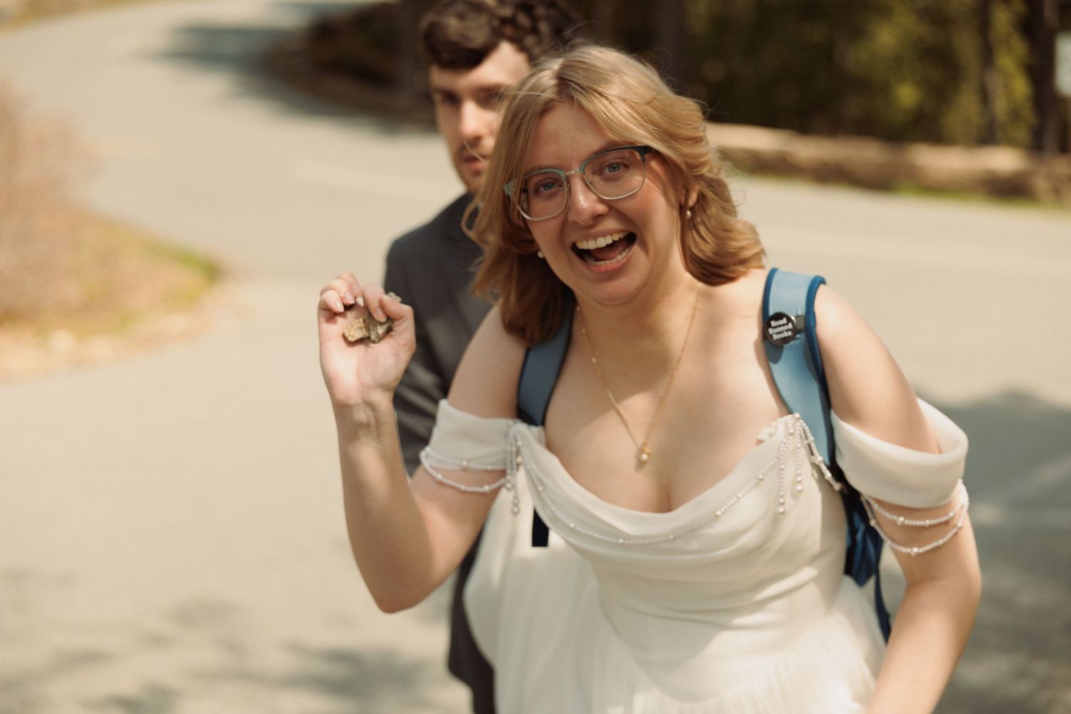 Smiling woman in white dress and glasses holding keys on sunny path with man in background.