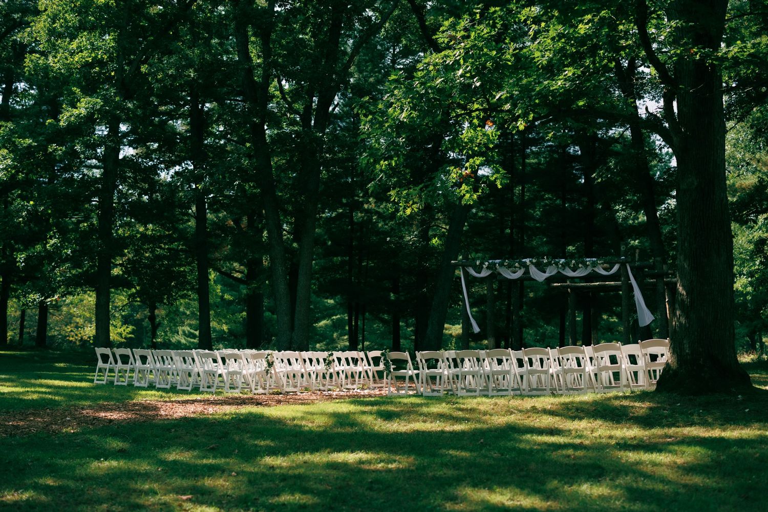 Panoramic view of empty white chairs set up for an outdoor wedding ceremony in a wooded area.