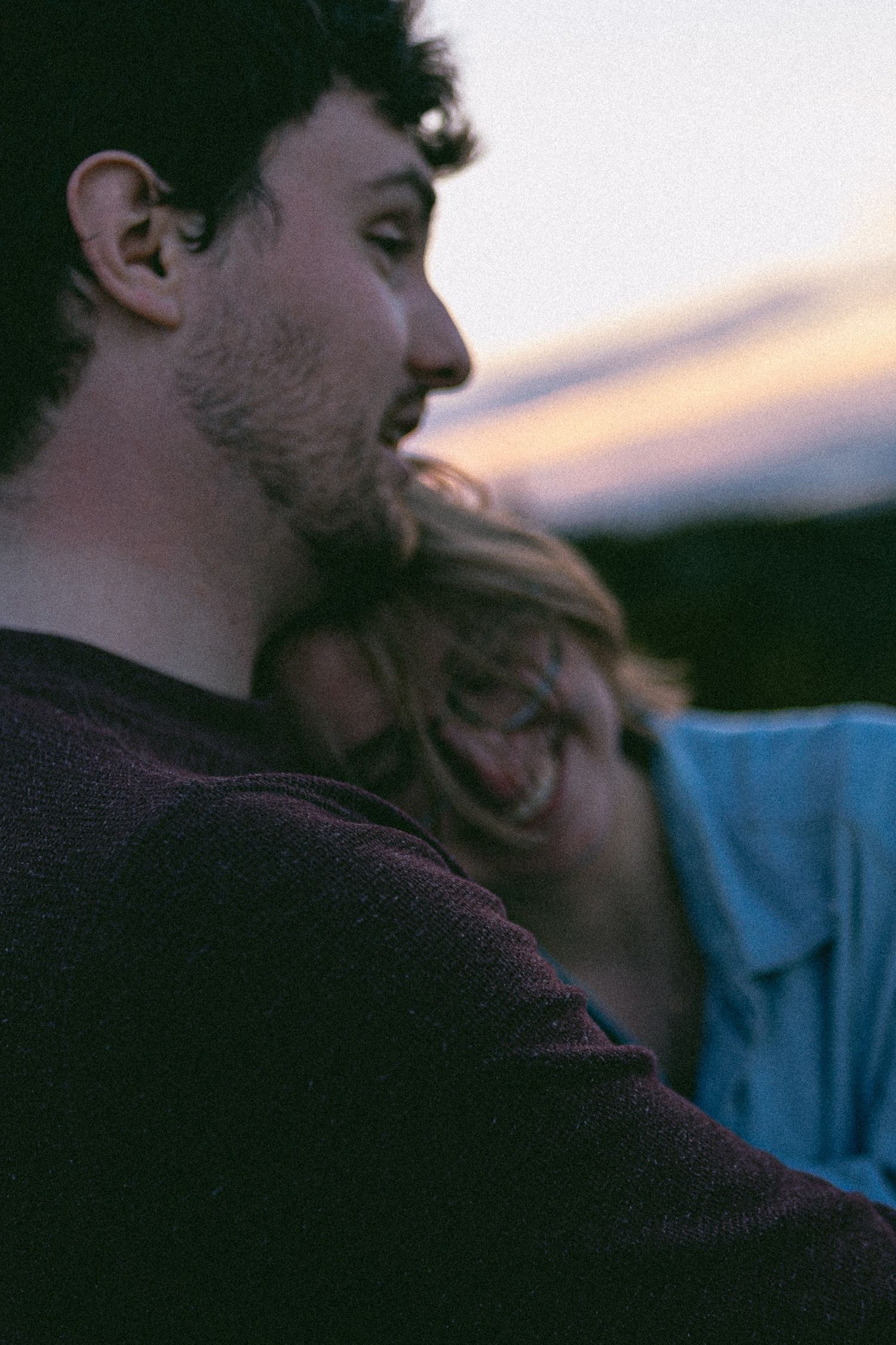 Couple sharing a tender moment at sunset, woman resting her head on man's shoulder in soft dusk light.