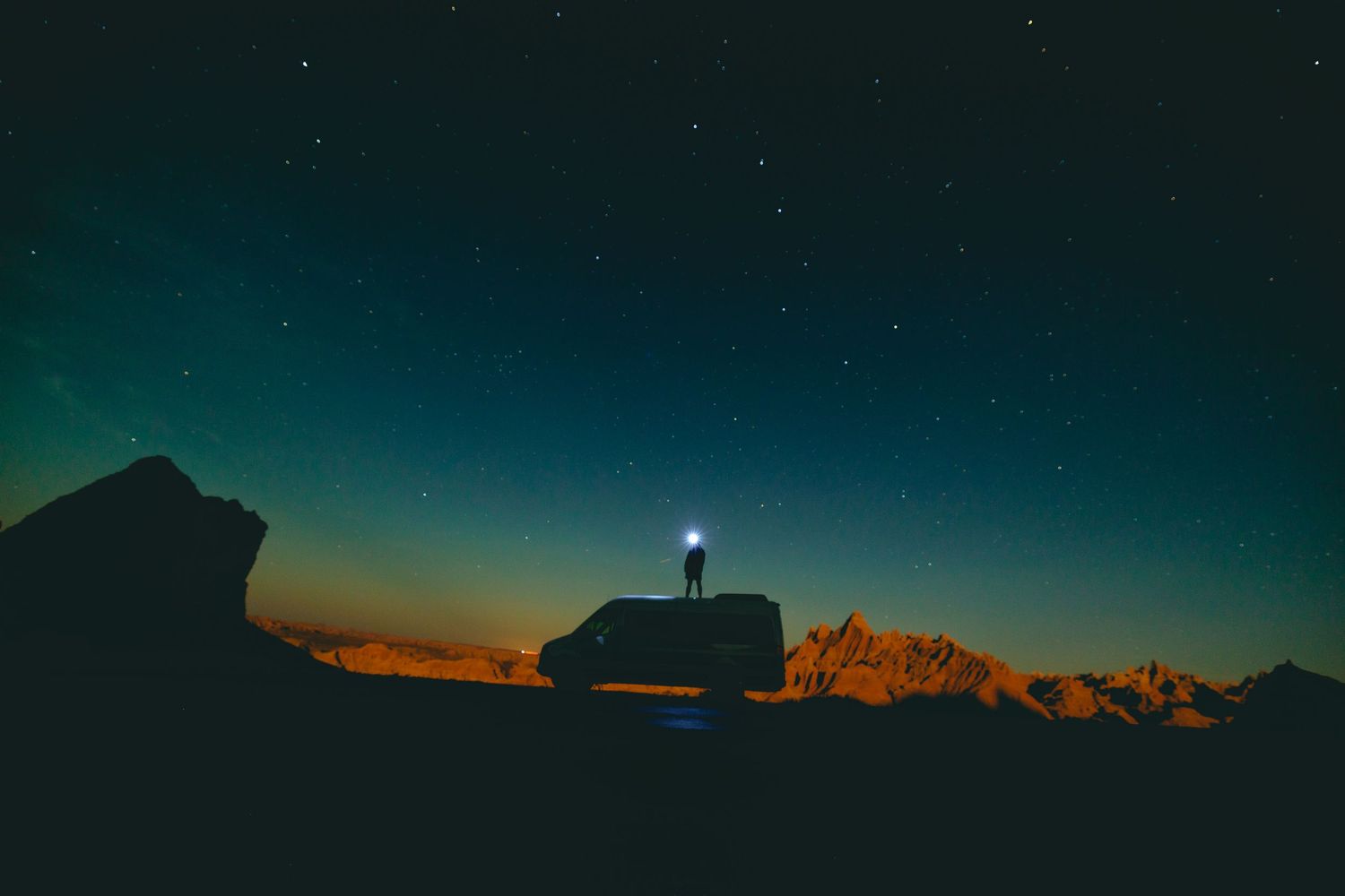 Starlit night sky over mountain silhouettes with a bright light on the horizon.